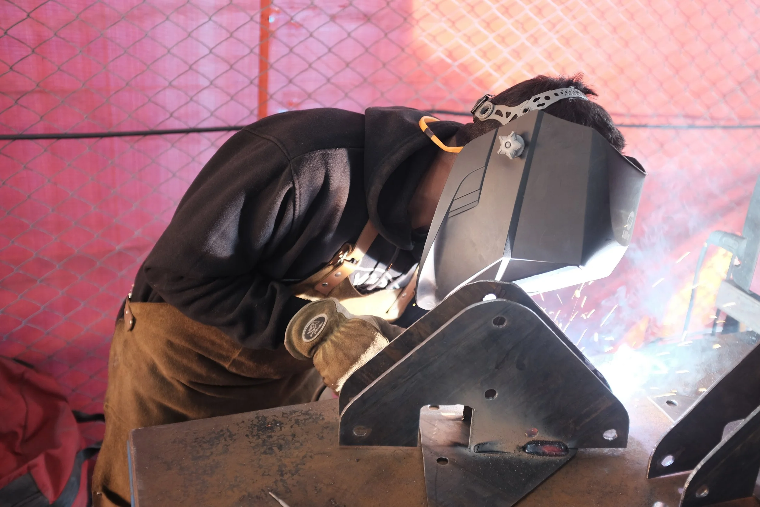 A person welding metal parts in a workshop, wearing a welding helmet, gloves, and a black jacket.