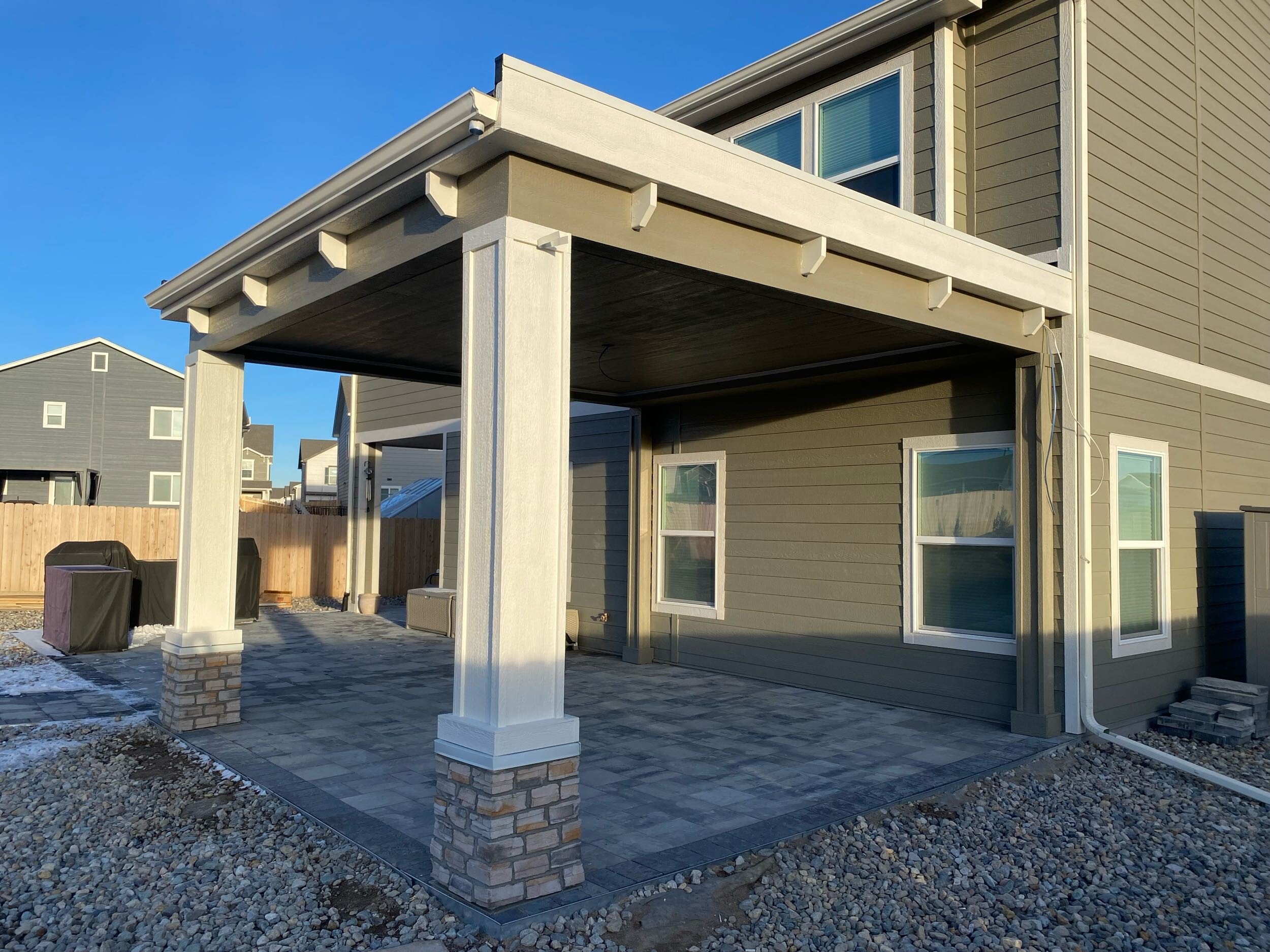 Newly constructed backyard patio with stone pavers, a covered veranda with white support columns, surrounded by a gravel yard, neighboring houses, and a wooden privacy fence.