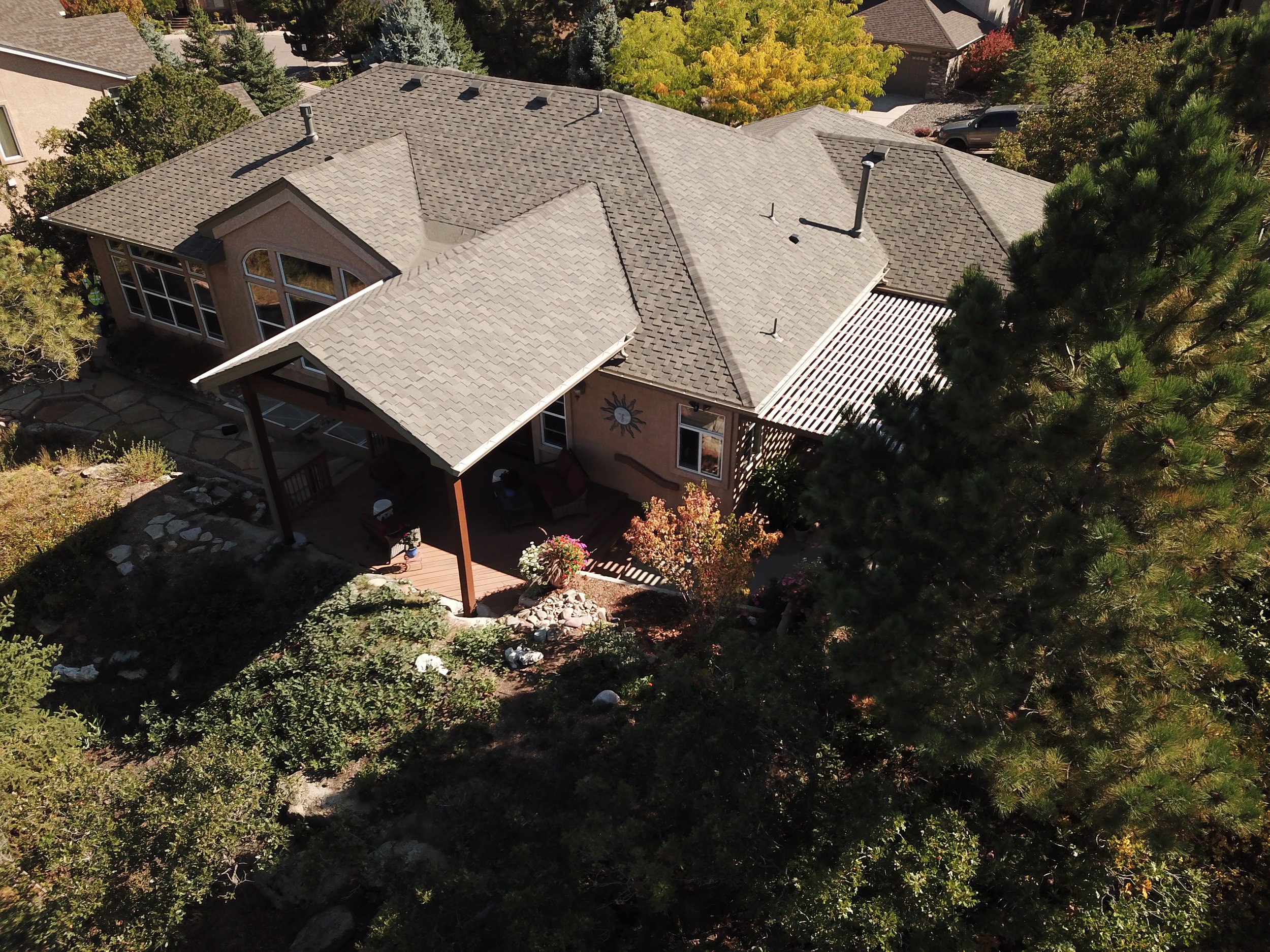 Aerial view of a suburban house with a gray shingle roof, a covered porch, and surrounding trees and shrubs.