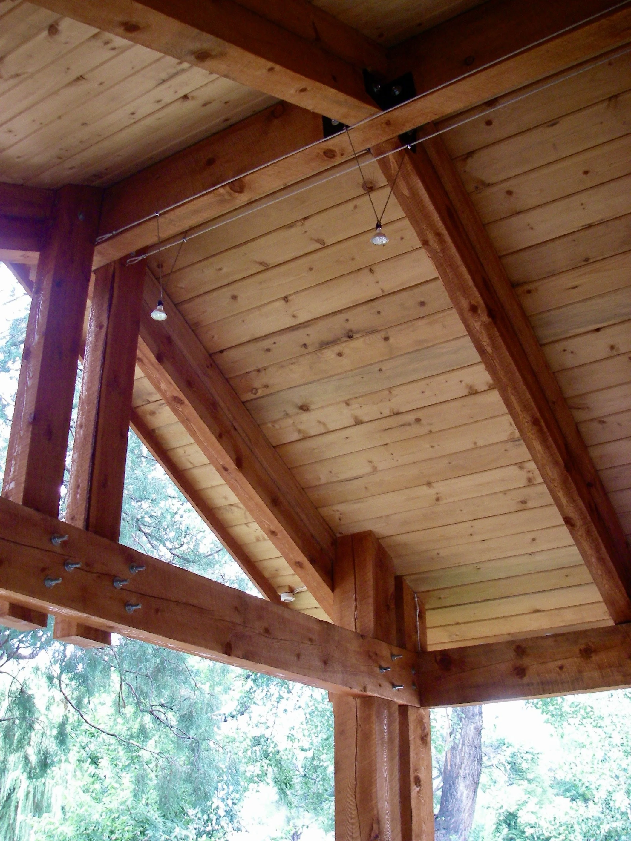 Wooden ceiling and beams of a porch or outdoor structure with string lights and trees in the background.