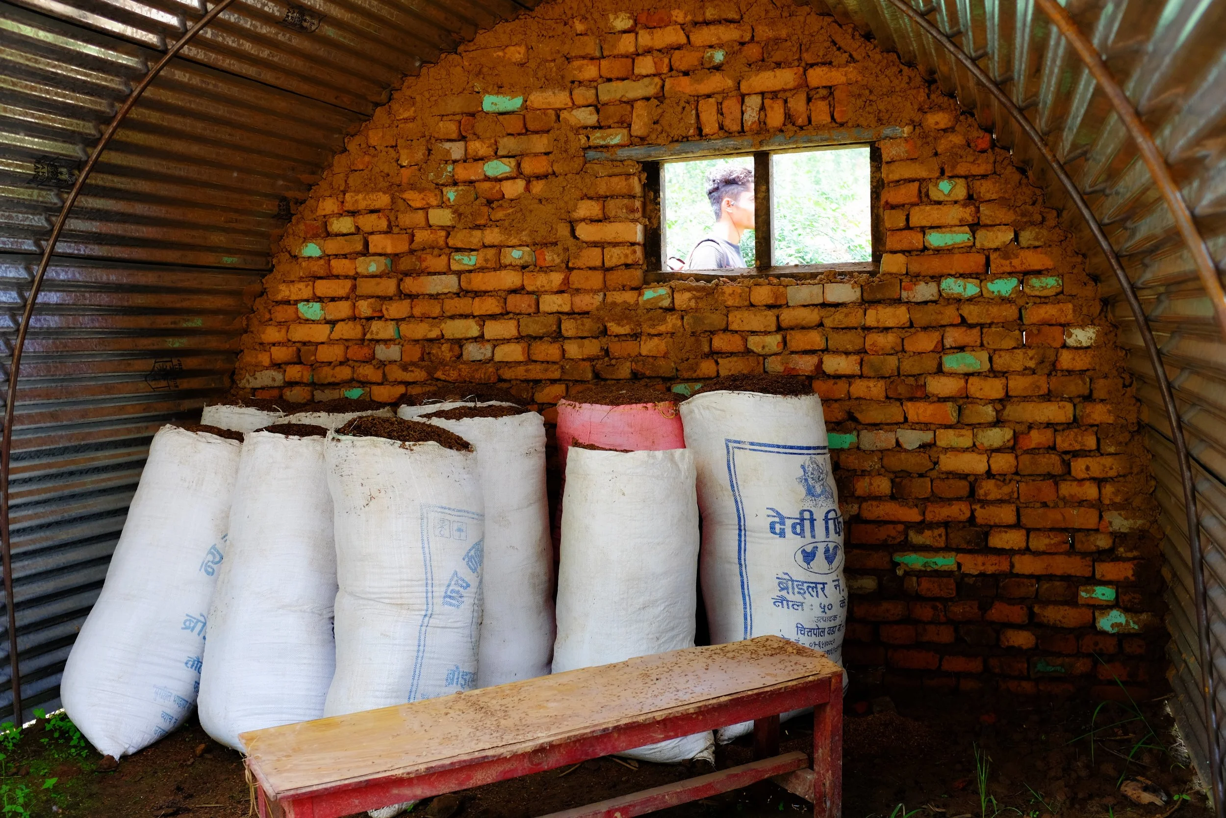 An unfinished brick wall inside a small, semi-circular shelter with a corrugated metal roof. Several white sacks filled with soil or compost are stacked against the wall. A small window cut out in the wall allows a person with glasses and a backpack 