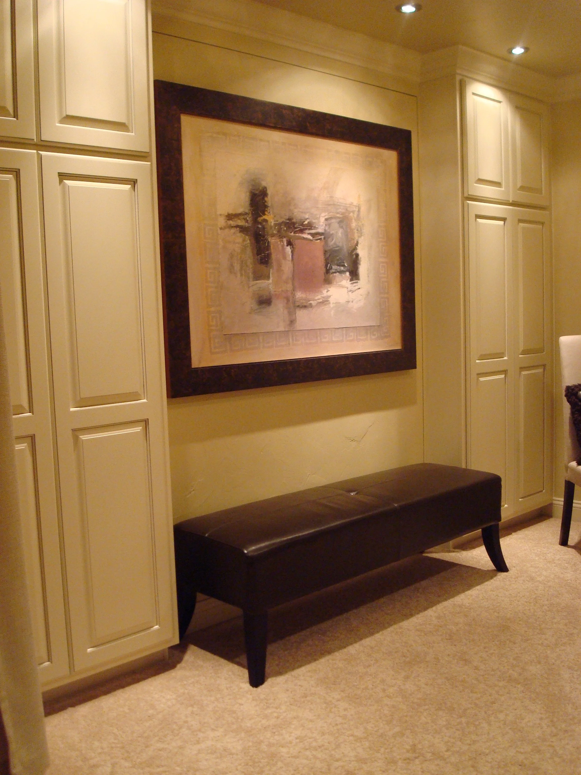 Interior room with beige cabinetry, a large abstract painting, a dark brown cushioned bench, and a beige carpeted floor.