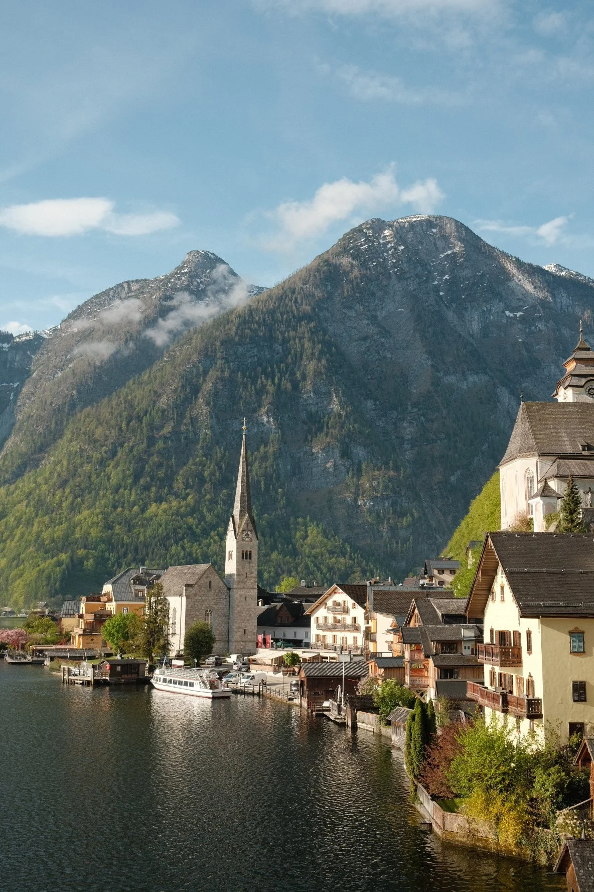 Scenic view of a lakeside village with a church and colorful houses, set against a backdrop of mountains and a partly cloudy sky.