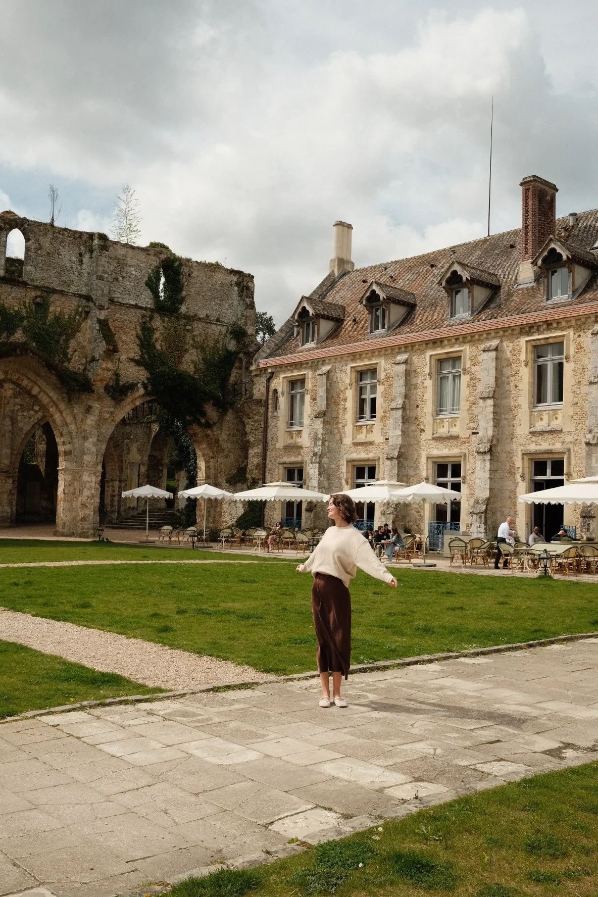 A woman in a cream sweater and brown skirt stands on a paved stone pathway with her arms outstretched, in front of an old stone building with outdoor tables and umbrellas, with a grassy area and ruins in the background.