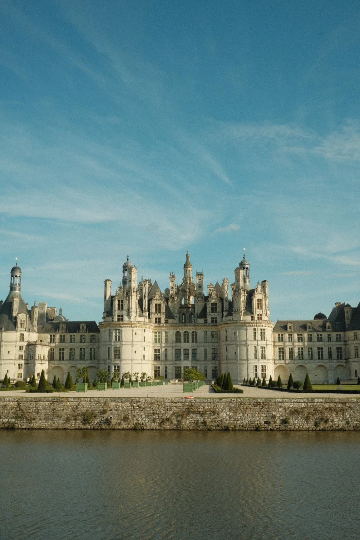 A large, ornate castle with turrets, towers, and intricate details, situated behind a stone wall and a body of water, under a clear blue sky.
