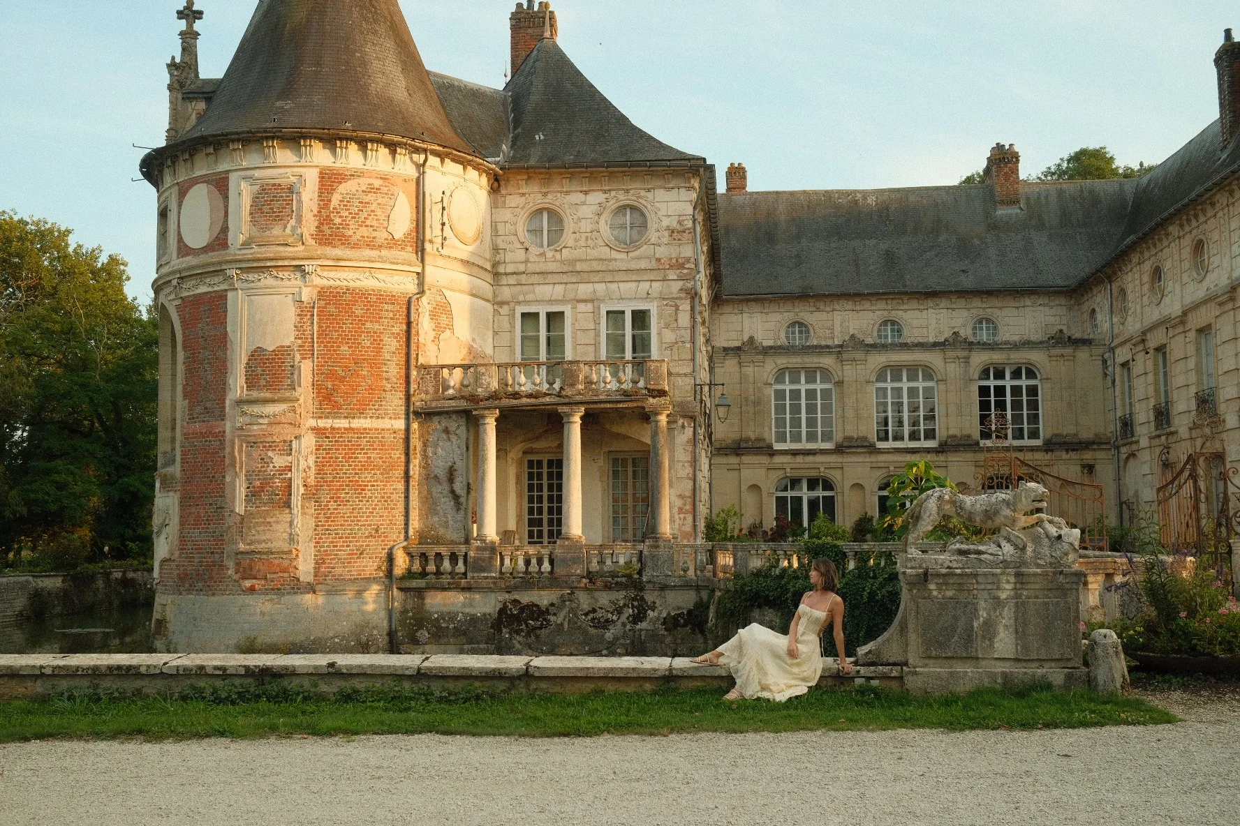 A woman in a long, white dress sits on a stone ledge in front of an old, ornate castle with a mix of brick and stone walls. The castle features a round tower, multiple windows, a small balcony, and a stone dragon sculpture on the right side. There ar