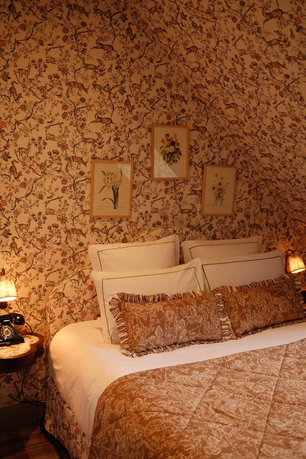 A cozy bedroom with floral-patterned wallpaper, a bed with white sheets and beige quilt, and multiple pillows including two large white ones with border and two smaller patterned brown ones. There are three framed floral artworks on the wall above the bed, two small bedside tables each with a vintage lamp and a rotary dial telephone.