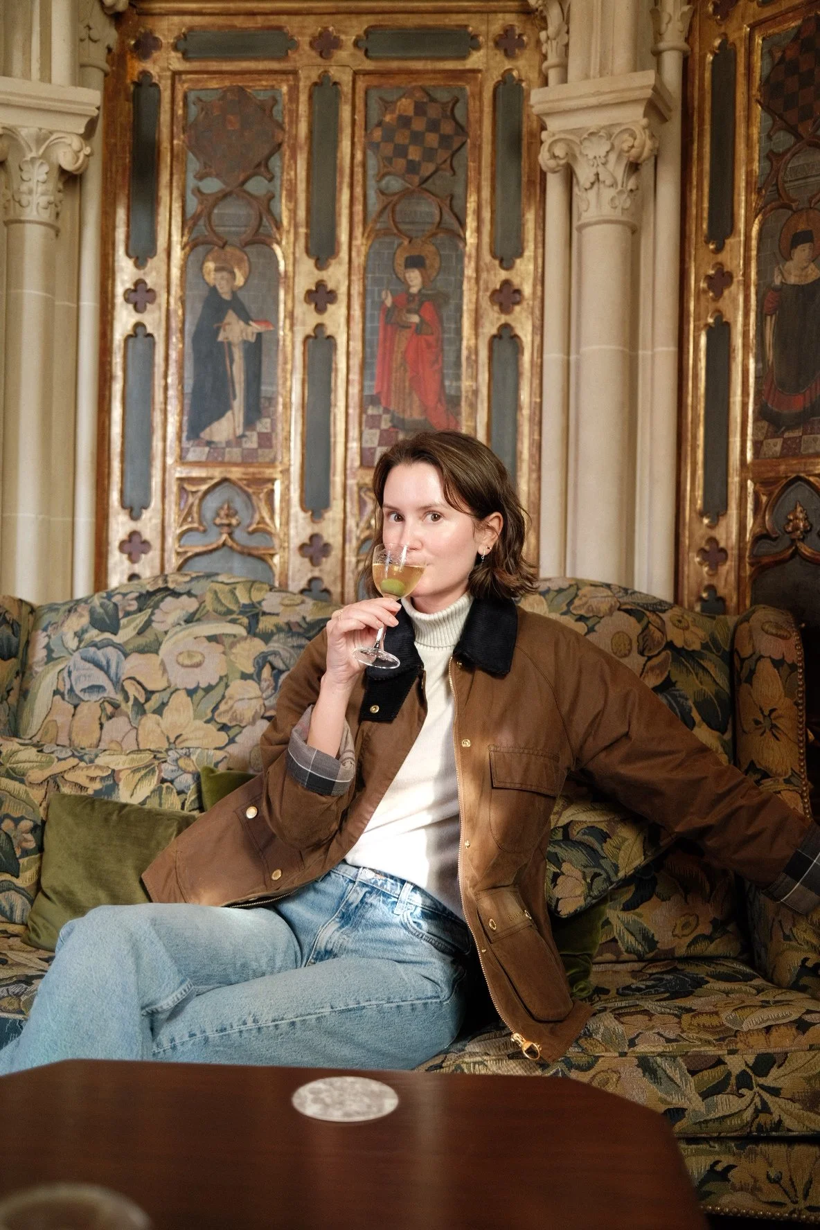 A woman sitting on a floral-patterned sofa, wearing a brown jacket, white turtleneck, and light blue jeans, holding a glass of white wine. She is in an ornately decorated room with historic paintings and columns in the background.