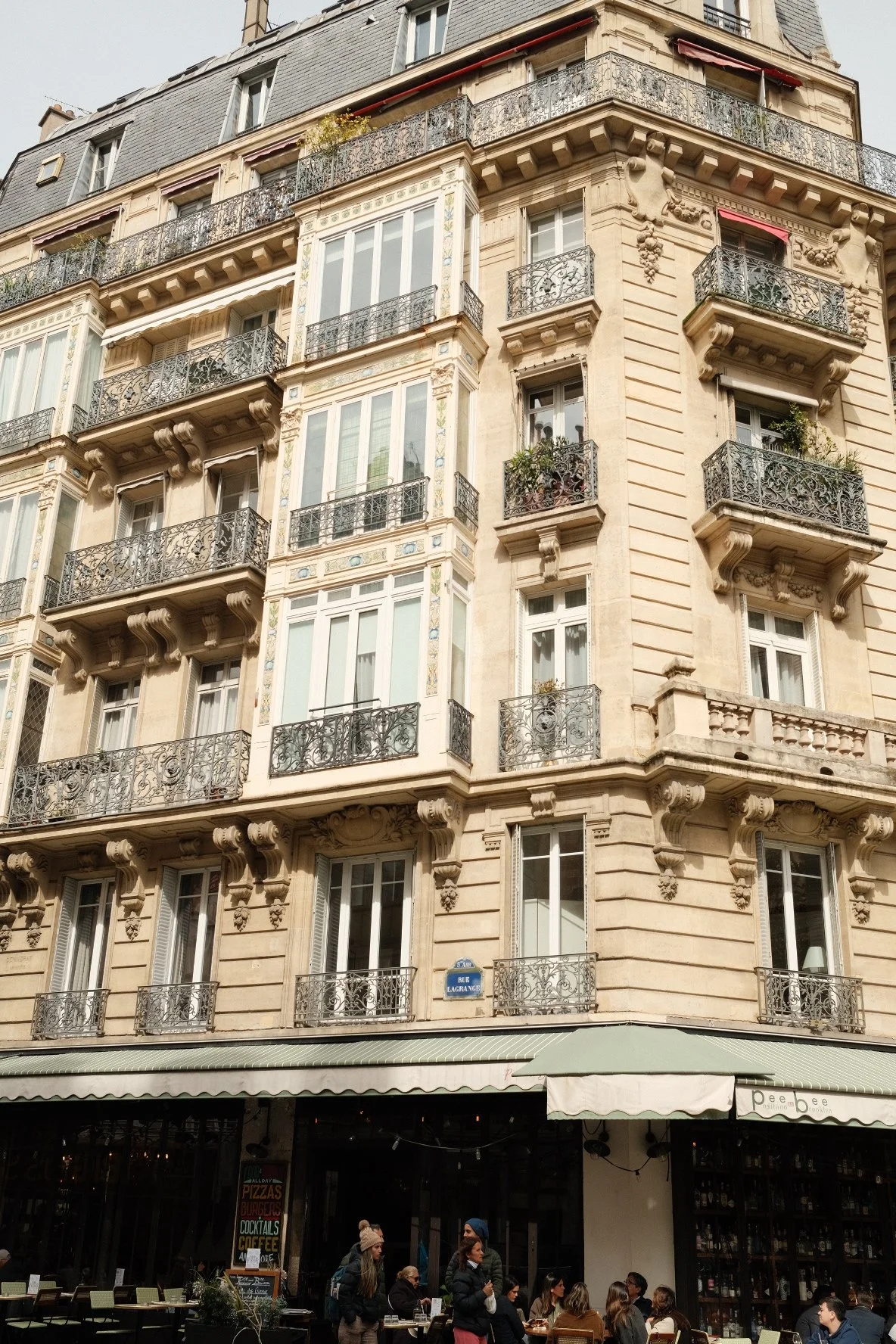 A multi-story Parisian-style building with ornate iron balcony railings and decorative stonework, located on Rue LaGrange, with a ground-floor café with outdoor seating and customers.