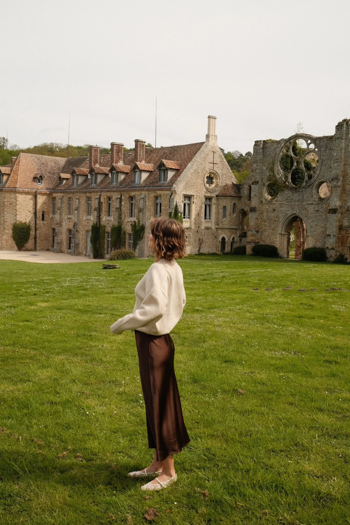 A woman with short curly hair, wearing a cream sweater, brown wide-leg pants, and white shoes, stands on a grassy field in front of an old stone building with large windows and a partially ruined section.