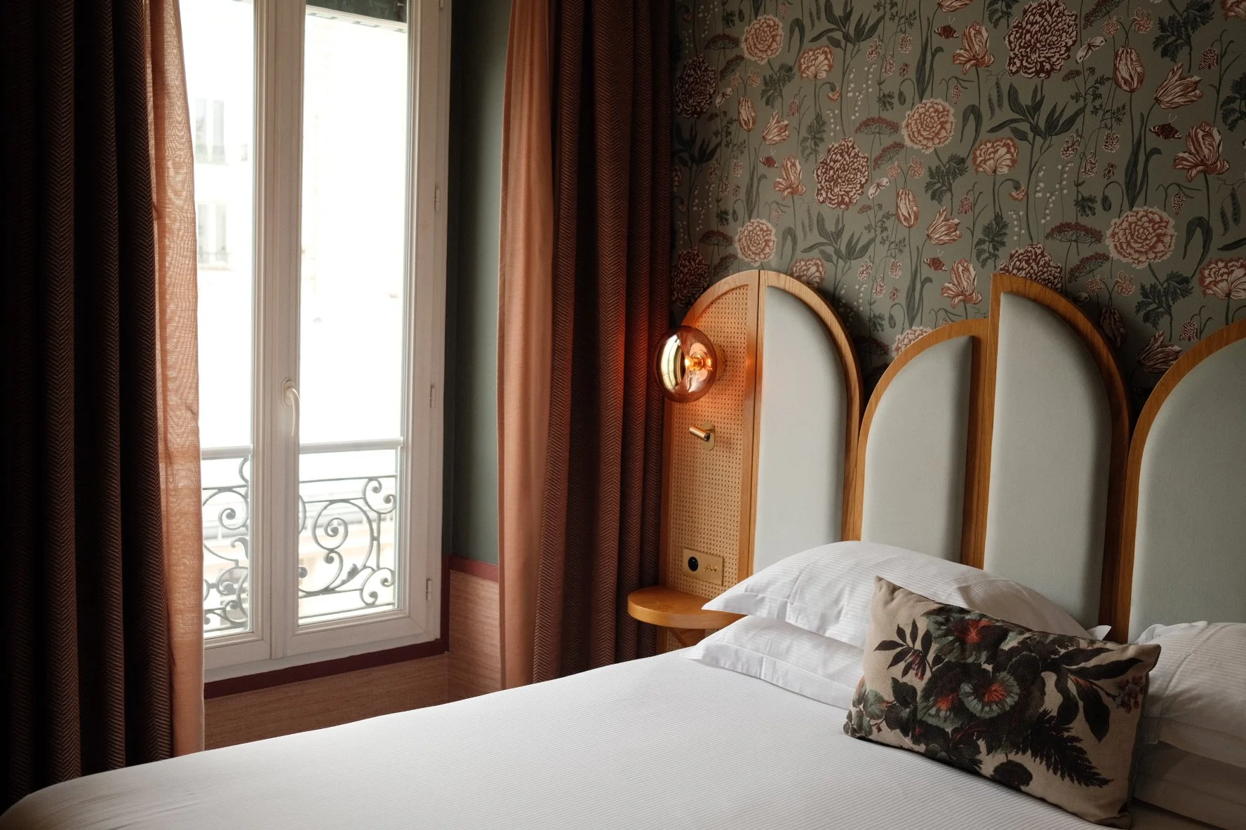 A hotel room with a bed, floral patterned pillow, bedside table, decorative headboard, floral wallpaper, and a large window with sheer curtains and an iron balcony railing visible outside.