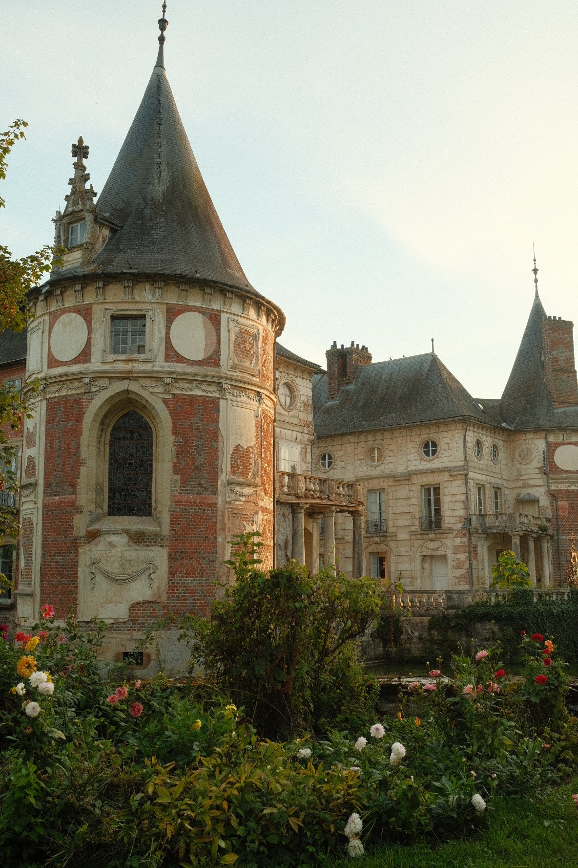 An old European castle with turrets, brick and stone walls, surrounded by colorful flowers and greenery.