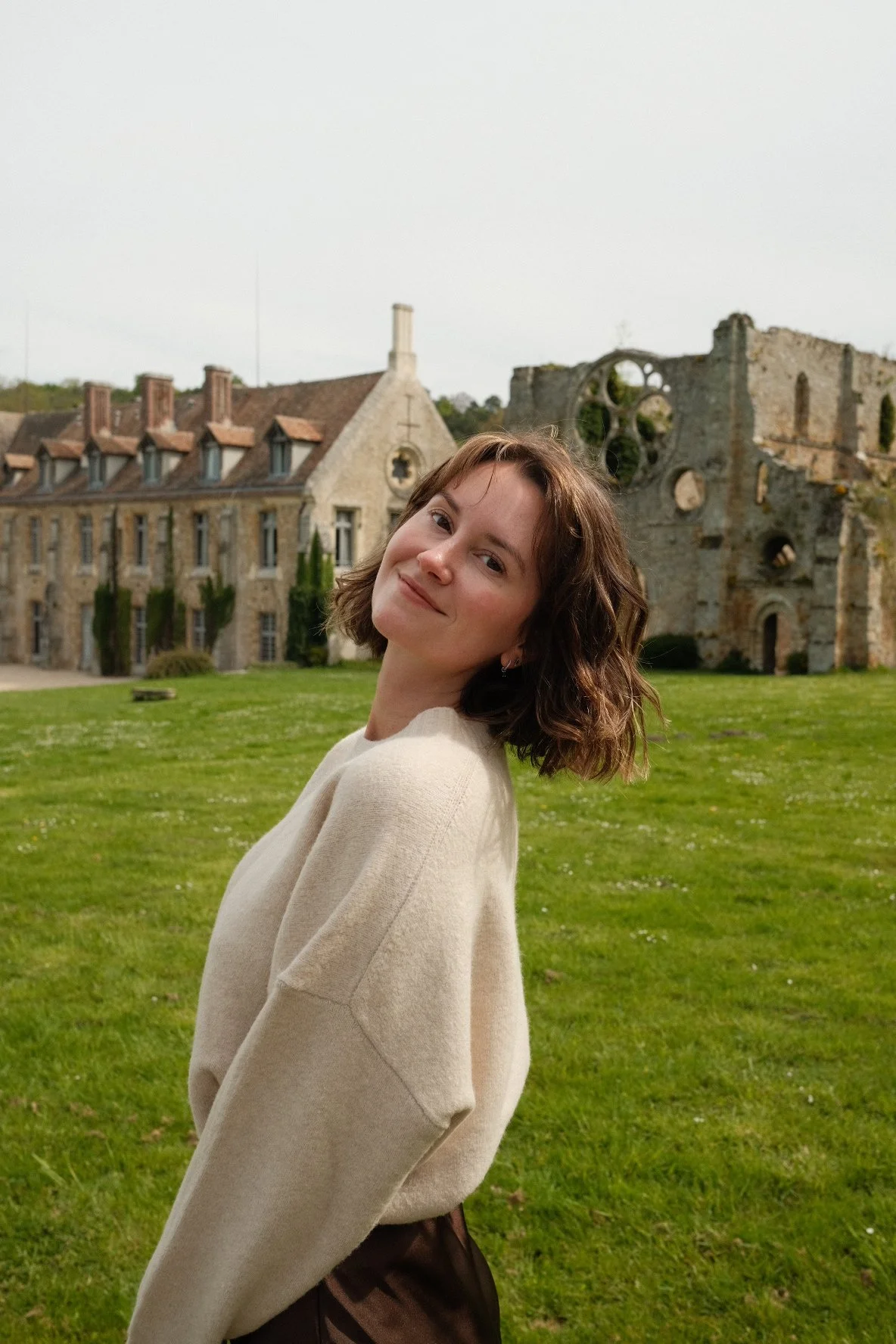 Young woman with short, wavy brown hair smiling at camera, standing on green grass in front of partially ruined stone structure and old stone building with chimney, overcast sky.
