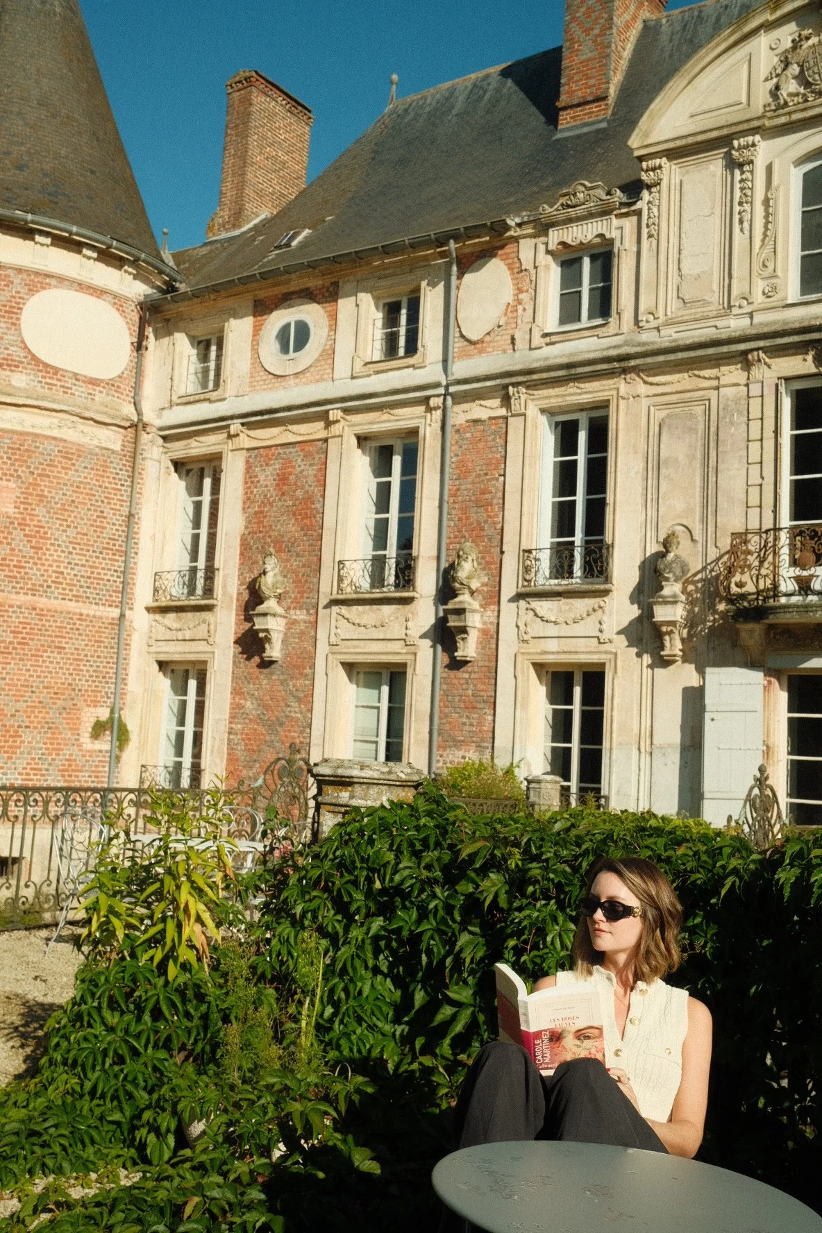 A woman with sunglasses sitting outdoors among green bushes, reading a book, with an old historic building in the background.