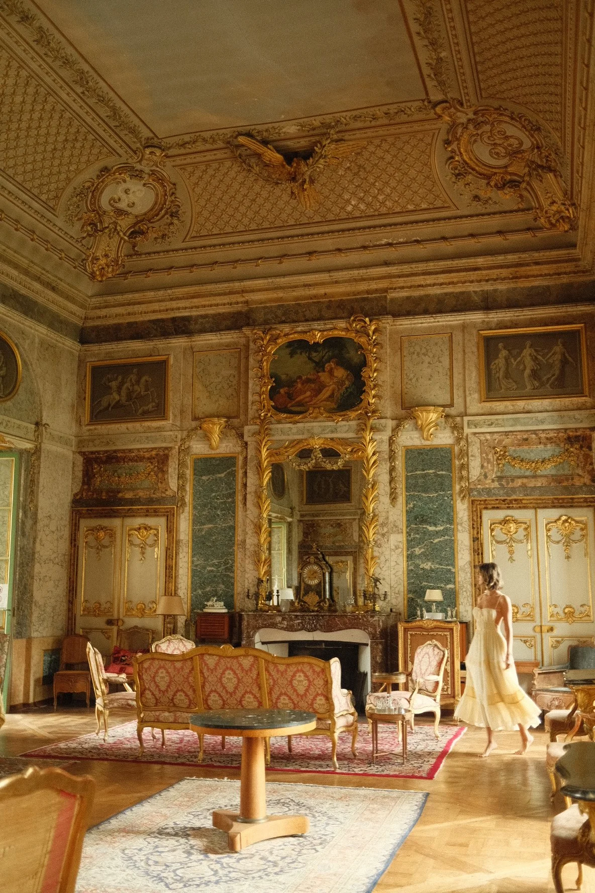 A woman in a yellow dress walking in a lavishly decorated room with ornate gold frames, green marble wall panels, classical paintings, vintage furniture, and a high ceiling with intricate gold molding.