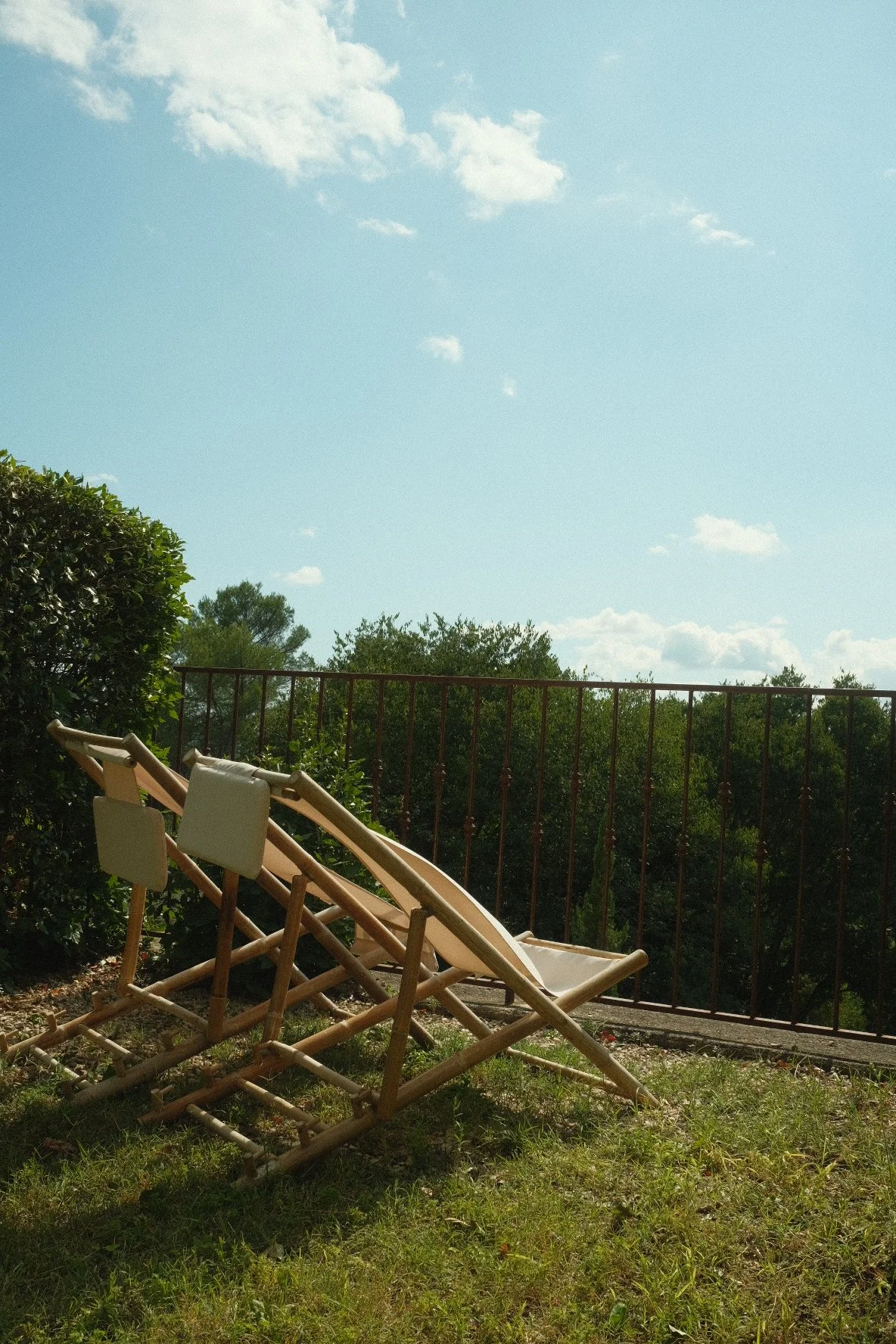 Three wooden deck chairs with white cushions are placed on a grassy area near a black metal railing, with lush green trees and a partly cloudy sky in the background.