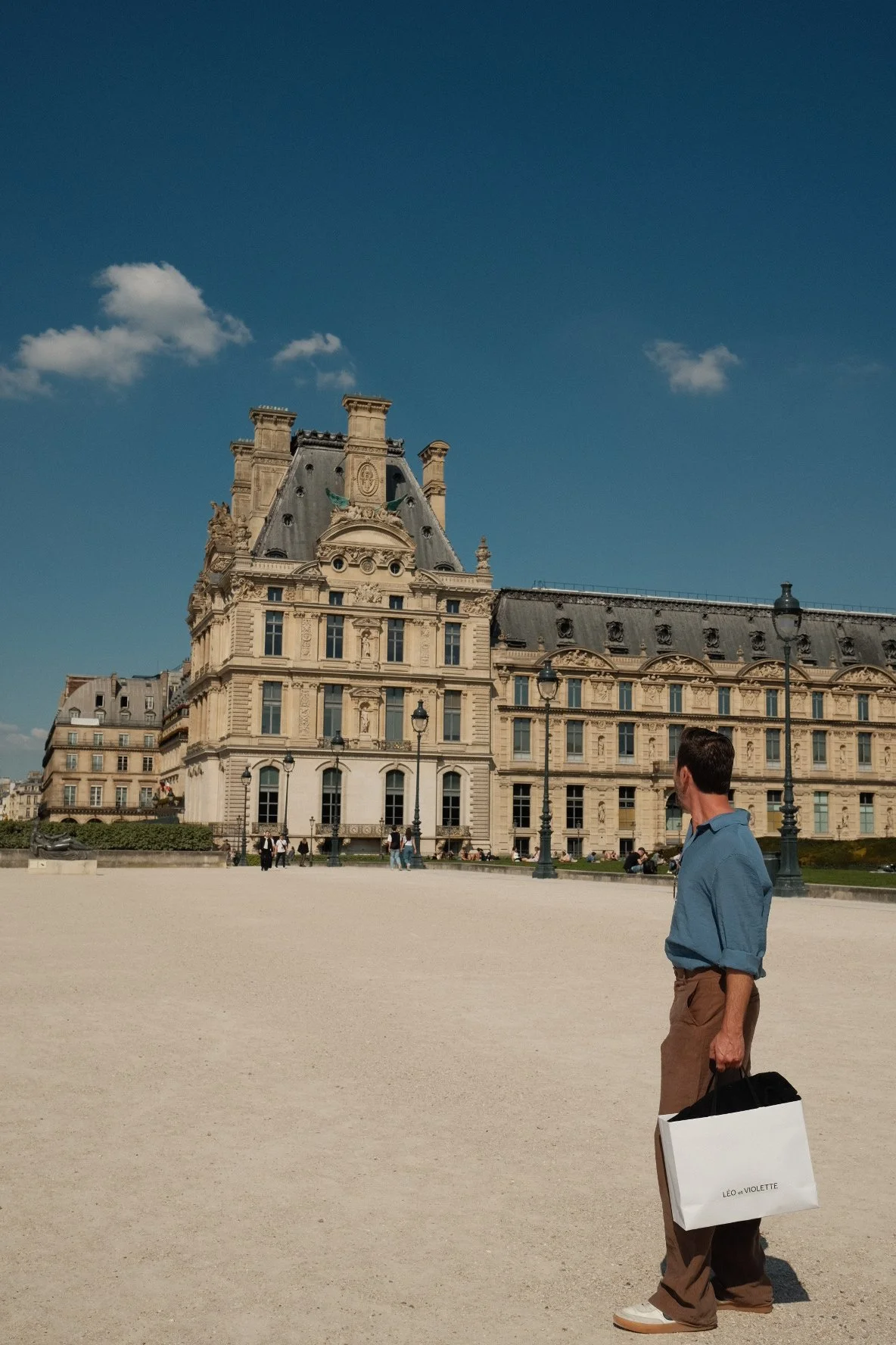 A man holding a shopping bag standing in front of a historic building with a clear blue sky and a few clouds.