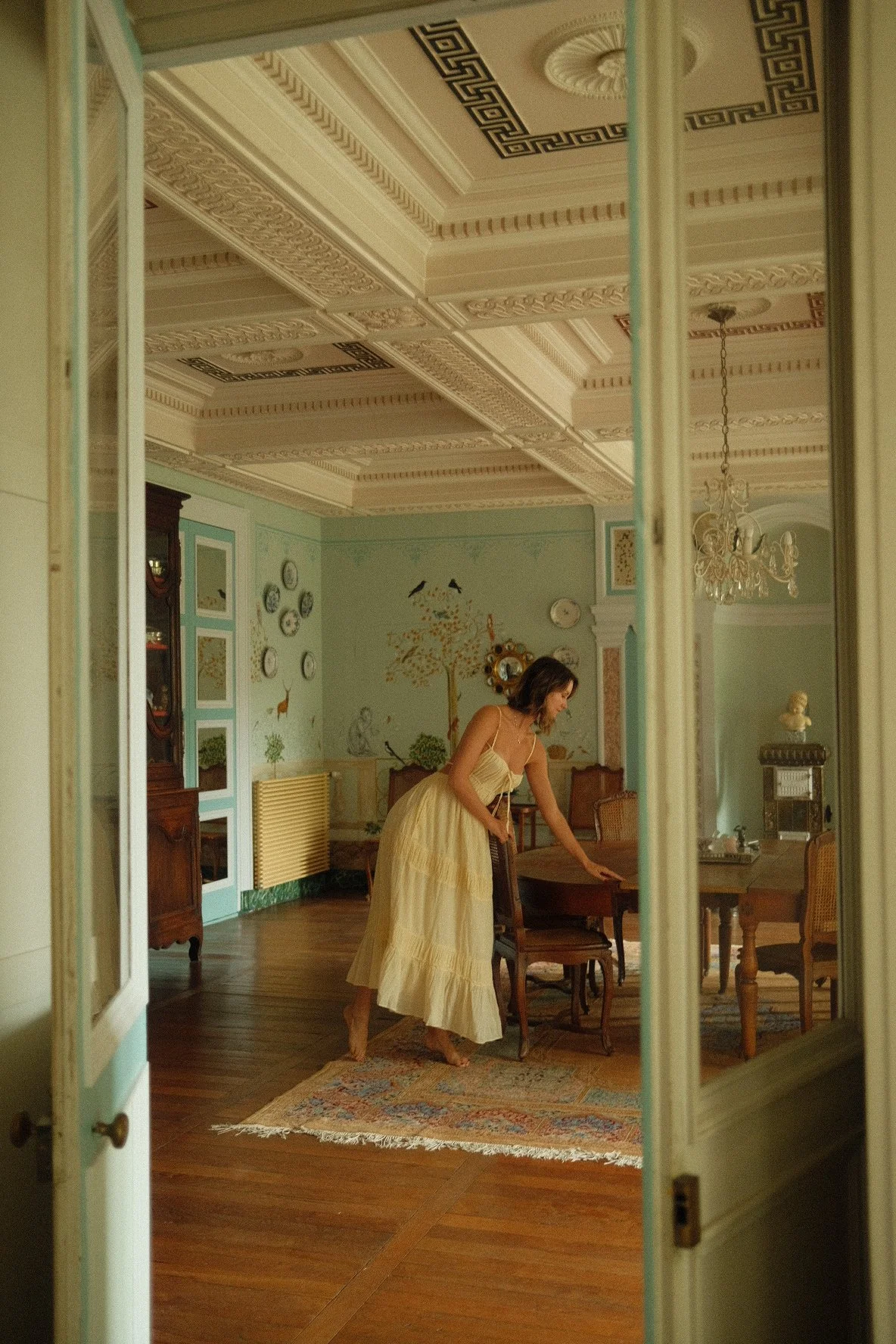 A woman in a cream-colored dress is standing on a colorful rug in a vintage dining room, leaning over a wooden table. The room features ornate ceiling moldings, a chandelier, ruffled curtains, and decorated walls with a tree and animal mural.