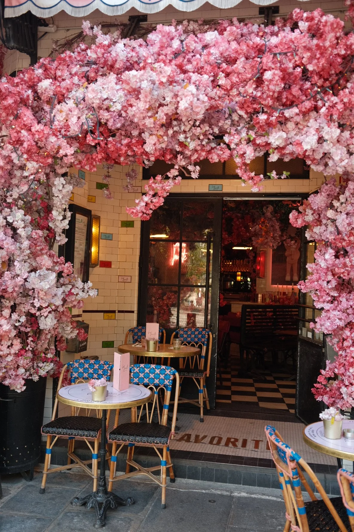 Café entrance decorated with pink cherry blossom flowers, outdoor seating with patterned chairs and small round tables, and a tiled floor with the word 'FAVORITE' near the entrance.