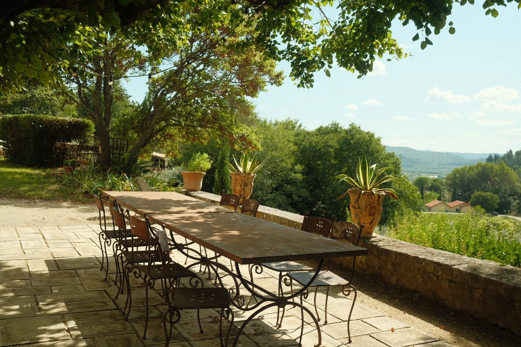 An outdoor patio with a long wooden table and metal chairs, surrounded by potted plants and lush green trees, overlooking a scenic landscape with houses and hills in the background on a sunny day.