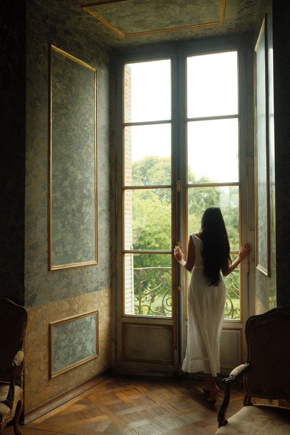 A woman in a white dress standing by an open window, looking outside at the greenery, in a vintage-styled room with ornate wall patterns and wooden parquet flooring.