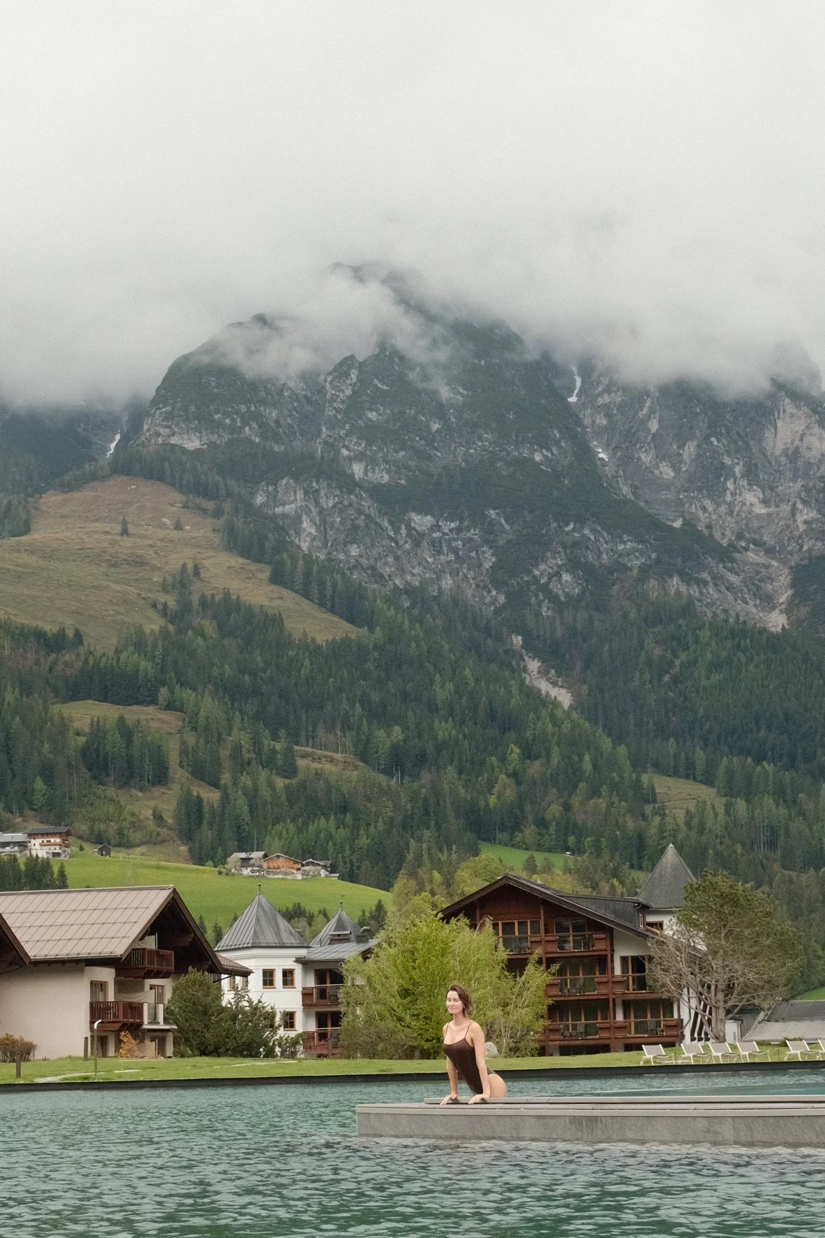 A woman in a swimsuit kneeling by a pool with a mountainous landscape and cloudy sky in the background.