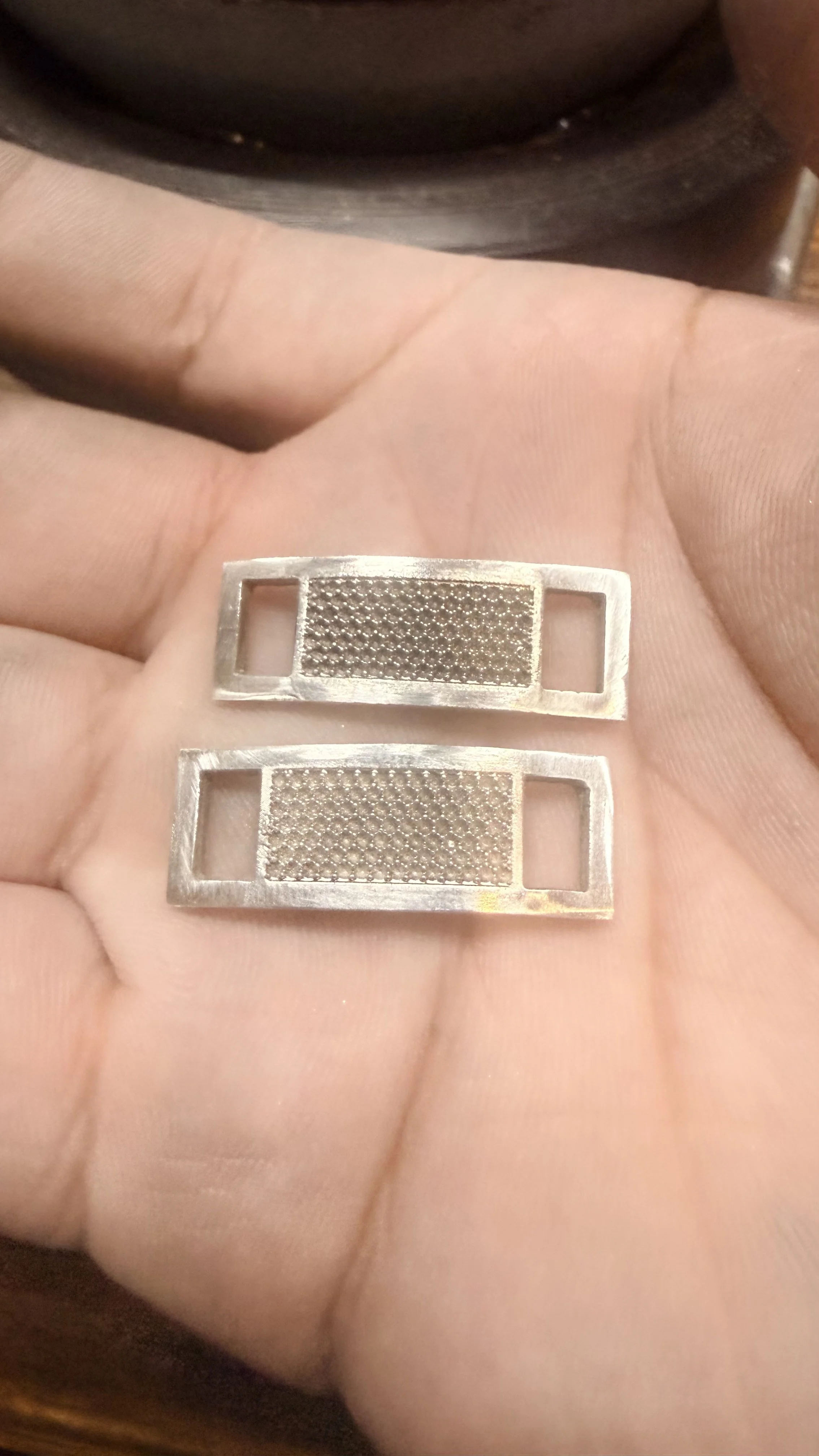 A person's hand holding two small metallic rectangular objects with a textured surface and open ends.