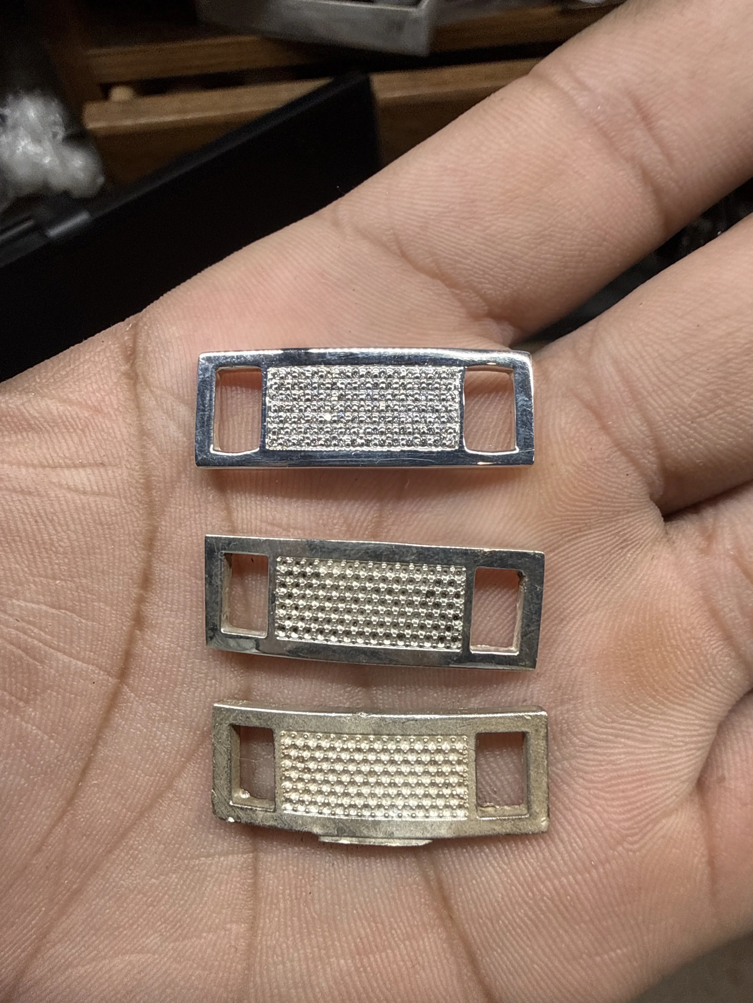 A hand holding three small rectangular metal objects with a textured pattern in the center and open slots on each side, possibly razor blade covers or decorative metal pieces.