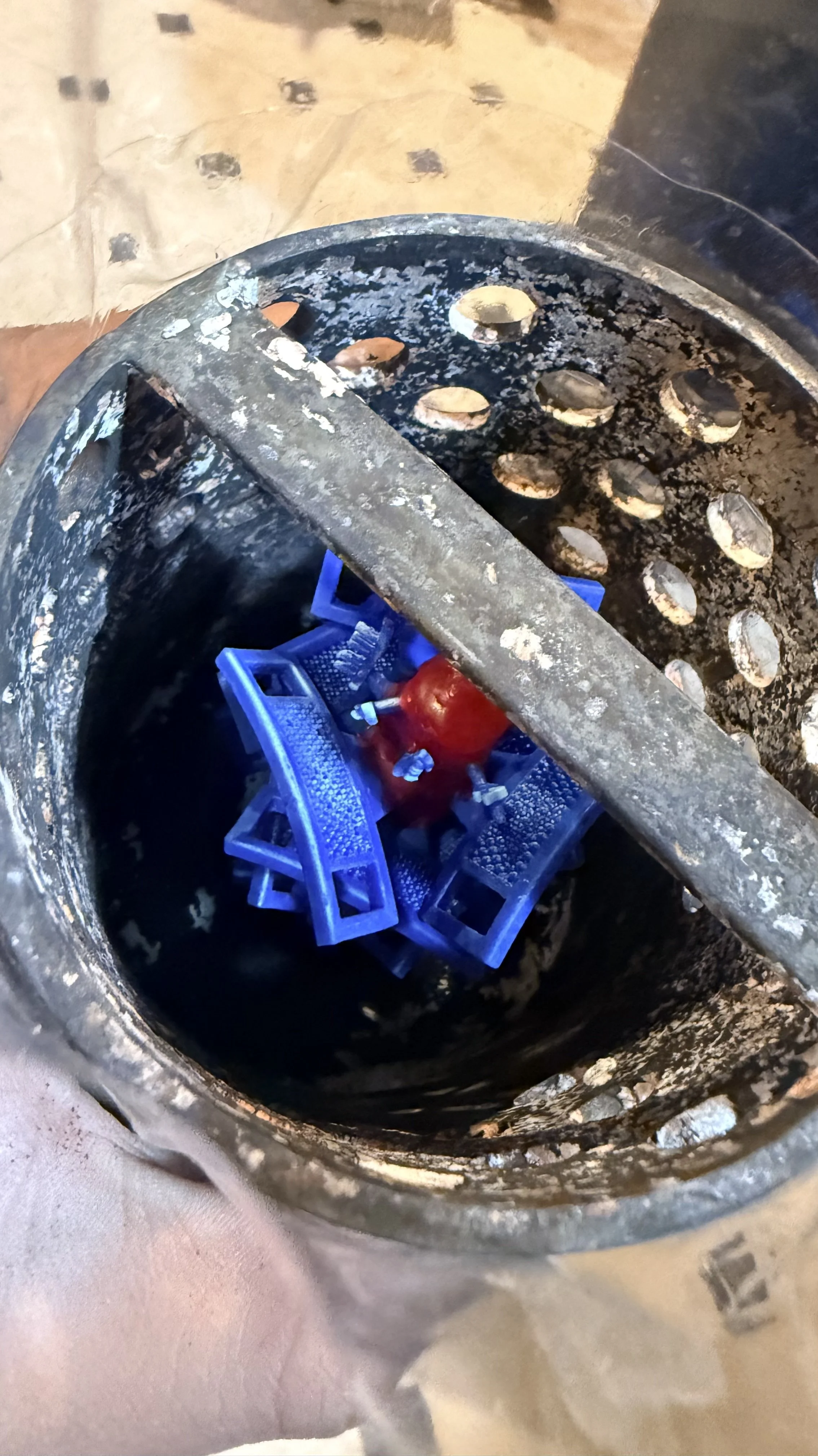 A rusty, black, cylindrical trash can with a metal bar across the top. Inside, there are discarded blue plastic flowerpot trays with a red apple placed on top of them.