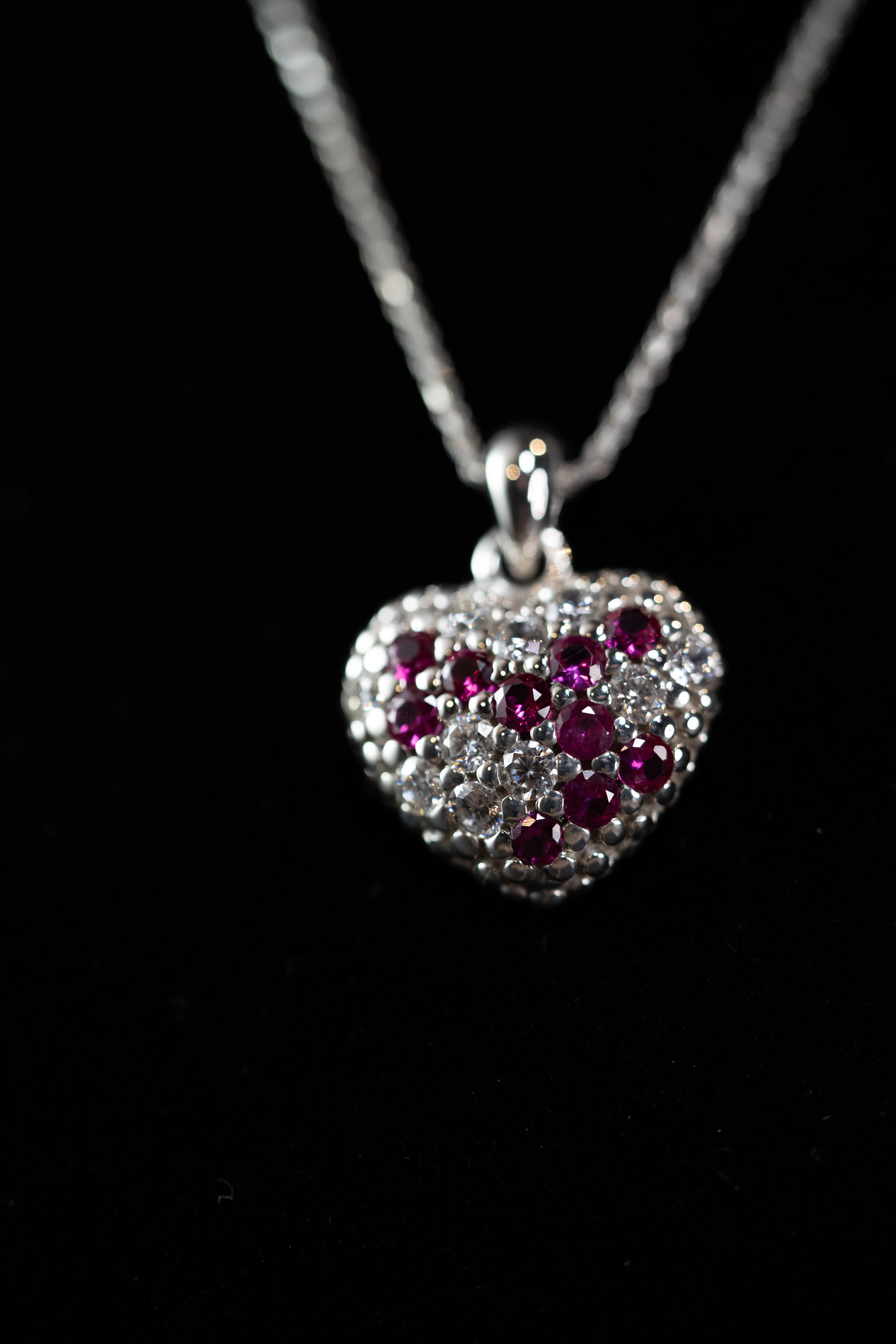 Silver heart pendant adorned with red and clear gemstones hanging on a silver chain against a black background.