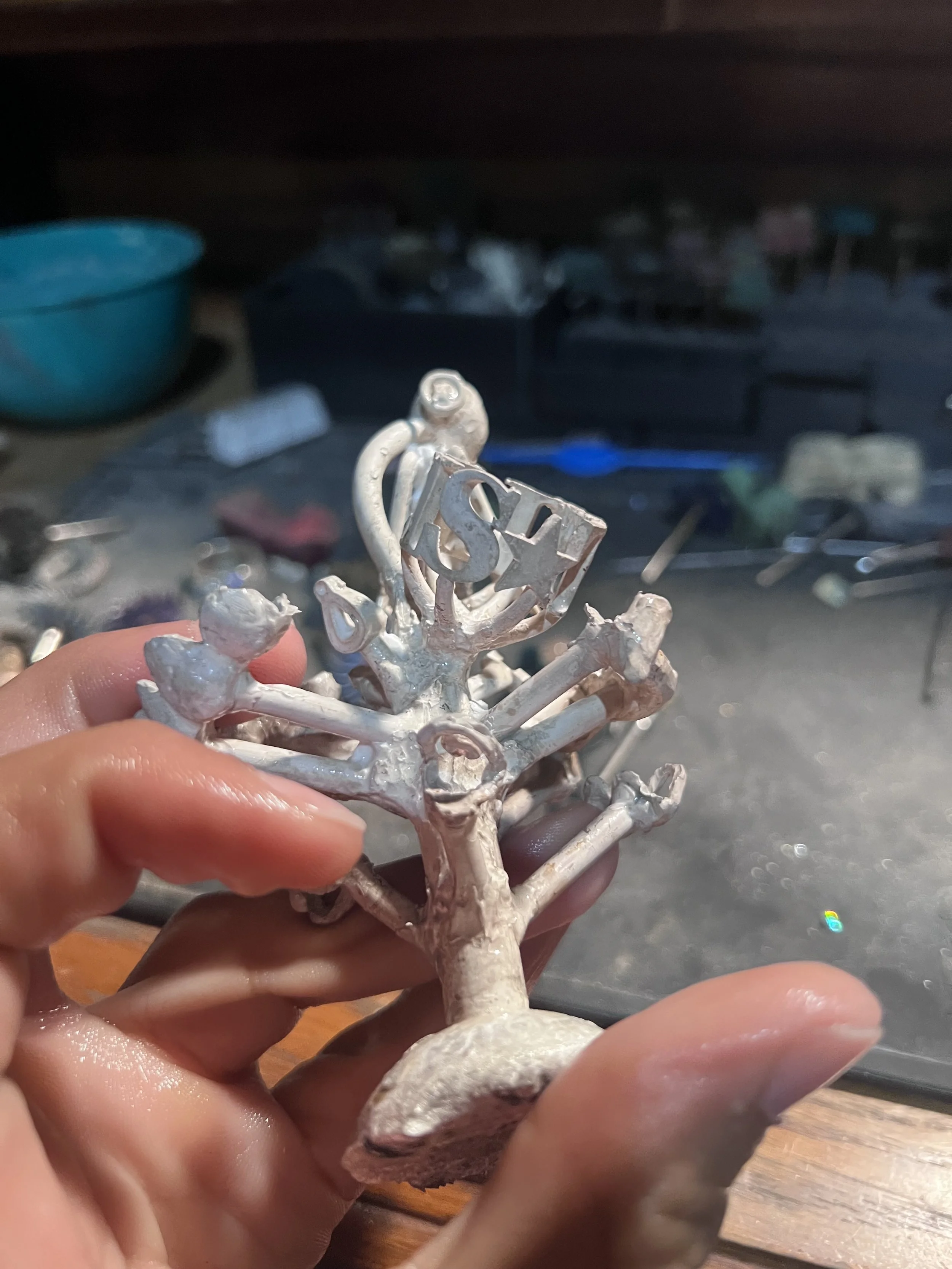 A person holding a small metal sculpture in front of a workbench with tools and supplies in the background.