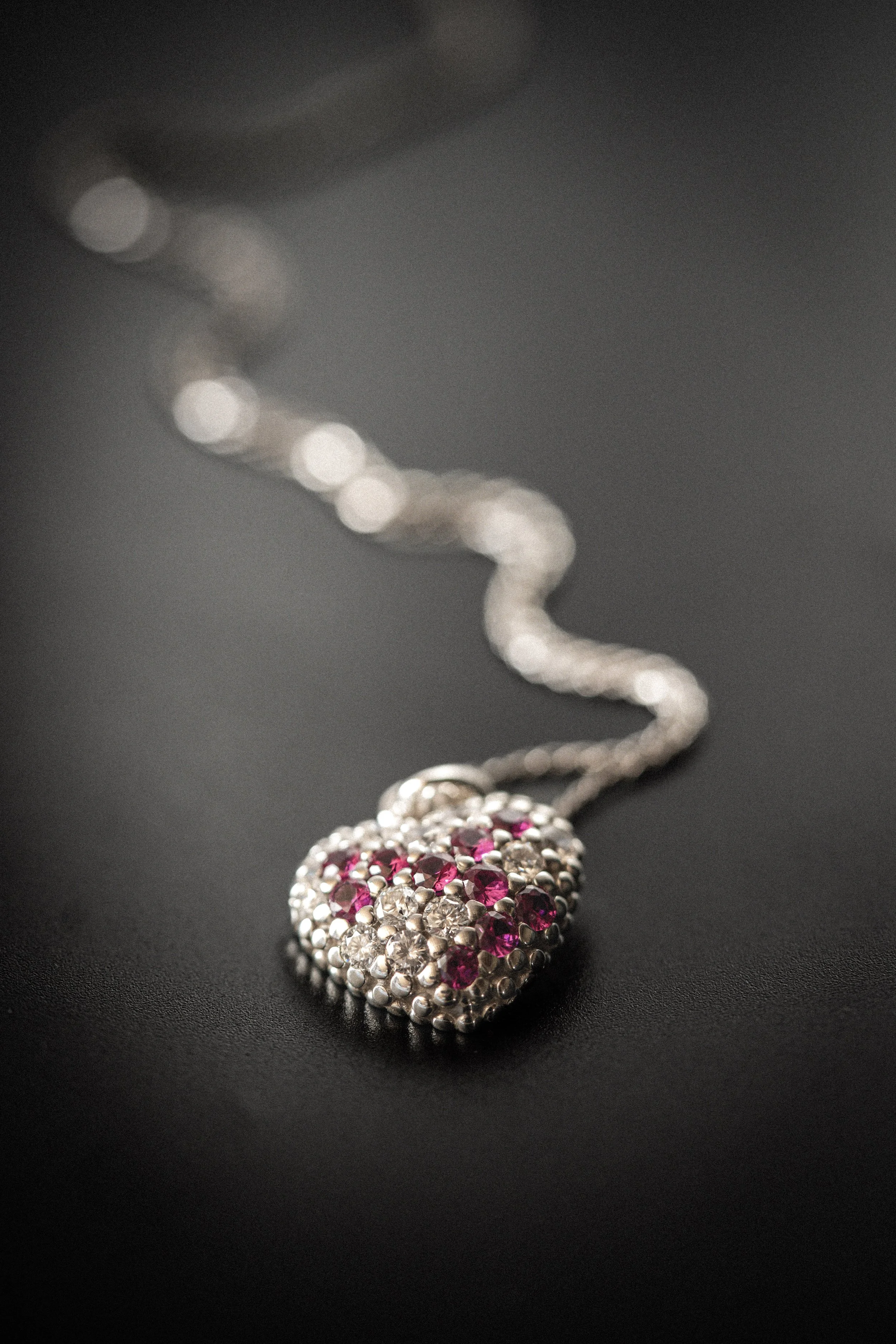 Close-up of a silver heart-shaped pendant necklace with pink and clear stones on a dark surface.