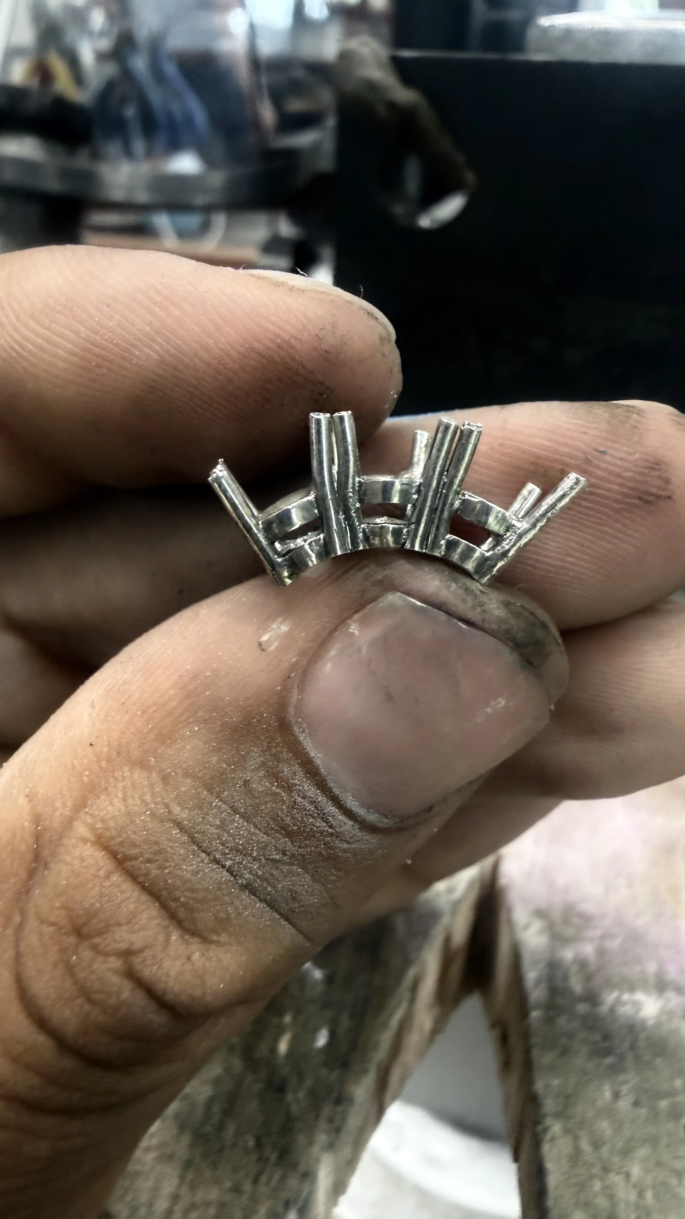 Close-up of a person's hand holding a metallic ring with an industrial design, showing vertical and curved elements, with a workshop background.