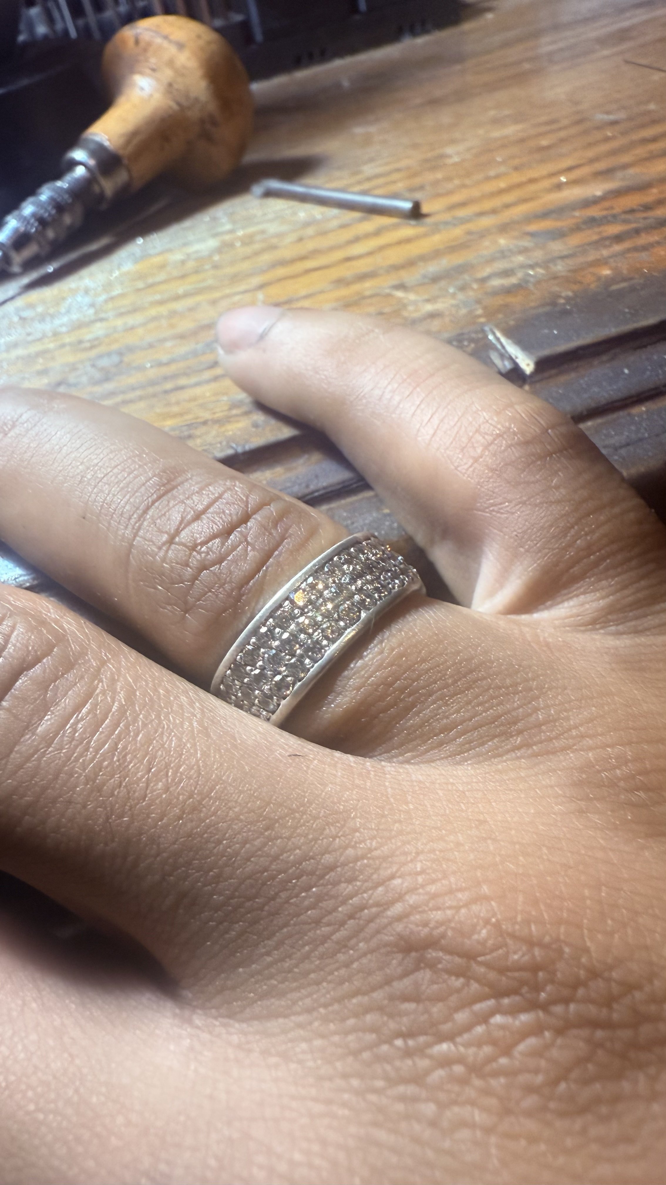 Close-up of a person’s hand wearing a diamond-studded ring, resting on a wooden workbench with jewelry tools in the background.