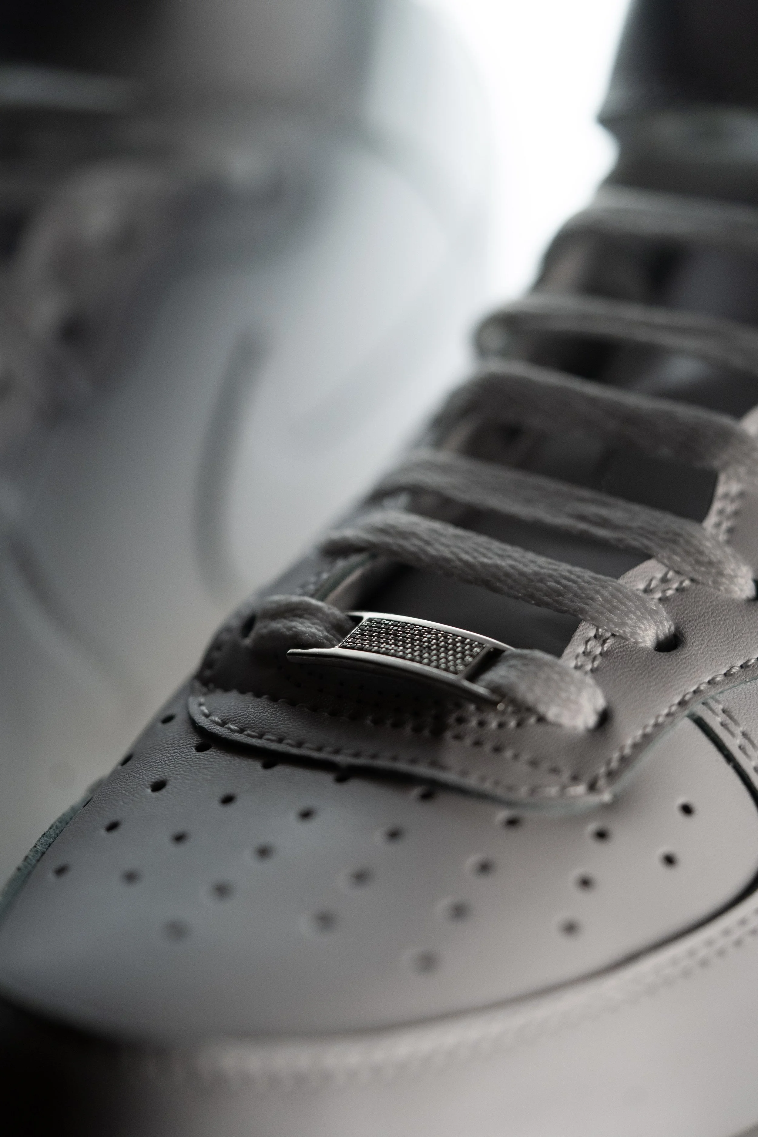 Close-up of a white sneaker with gray laces and metal lace holder, showing detailed stitching and perforations on the toe cap.