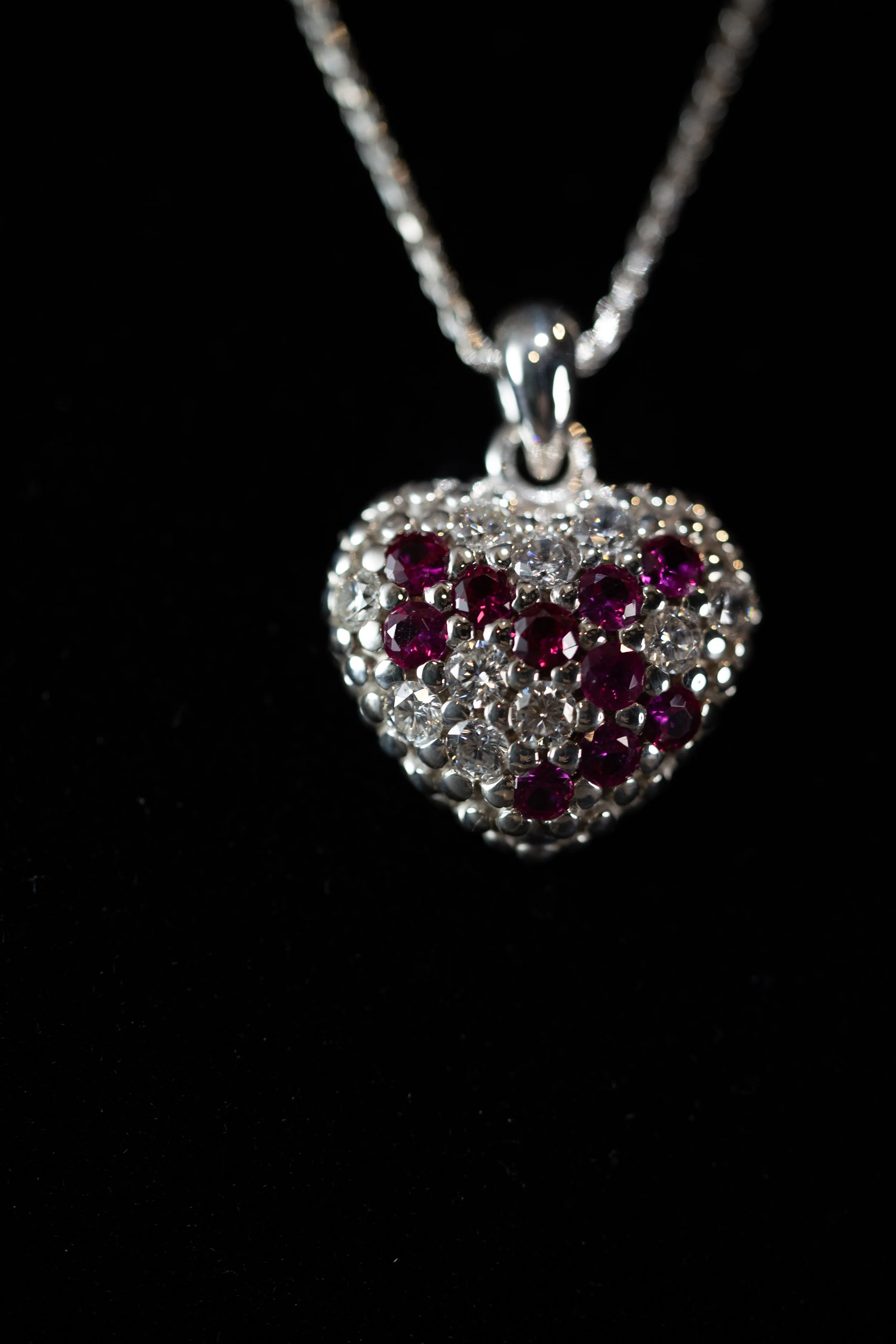 A silver heart-shaped pendant necklace with red and clear gemstones on a black background.