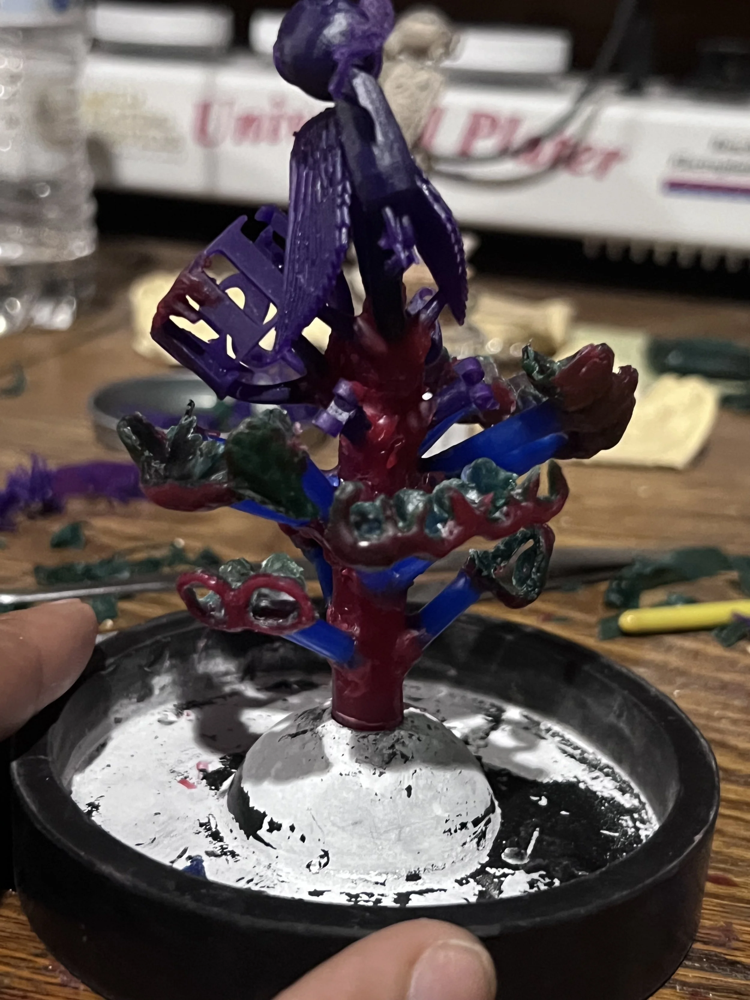 A small, colorful, handmade tree sculpture made of plastic and painted in purple, blue, green, and red, mounted on a white base inside a black container.