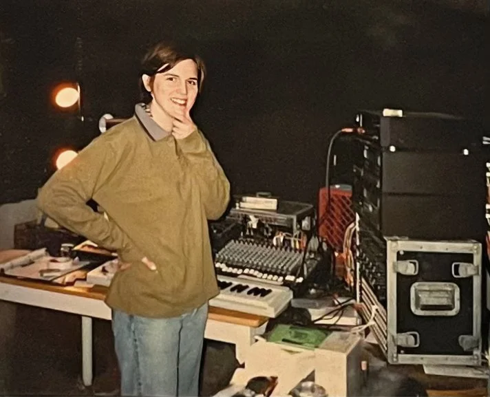 A woman with short brown hair wearing a tan jacket stands in a music or DJ studio with equipment, smiling and touching her chin.