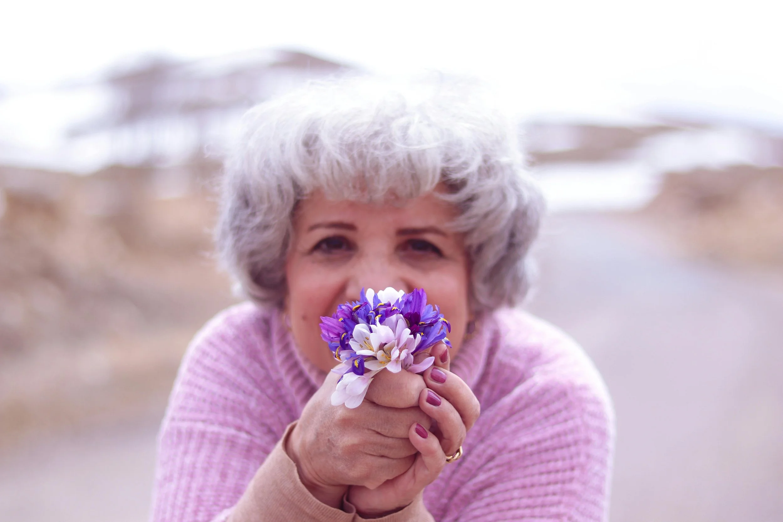 An elderly woman with curly gray hair and a pink sweater holding a bouquet of purple and white flowers near her face outdoors.