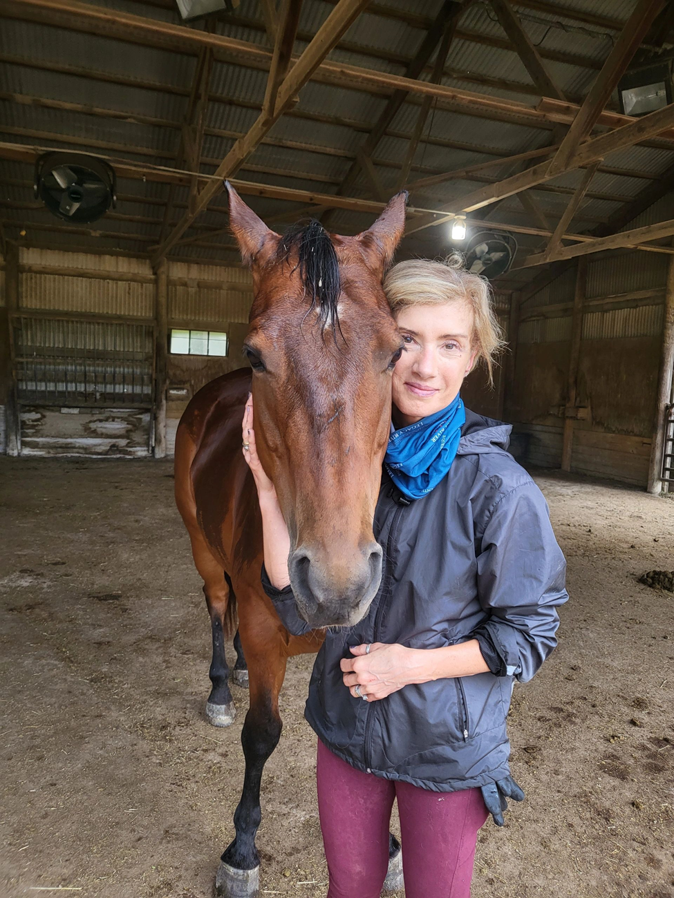 A woman with blonde hair and a gray jacket standing next to a brown horse with a black mane in a barn.