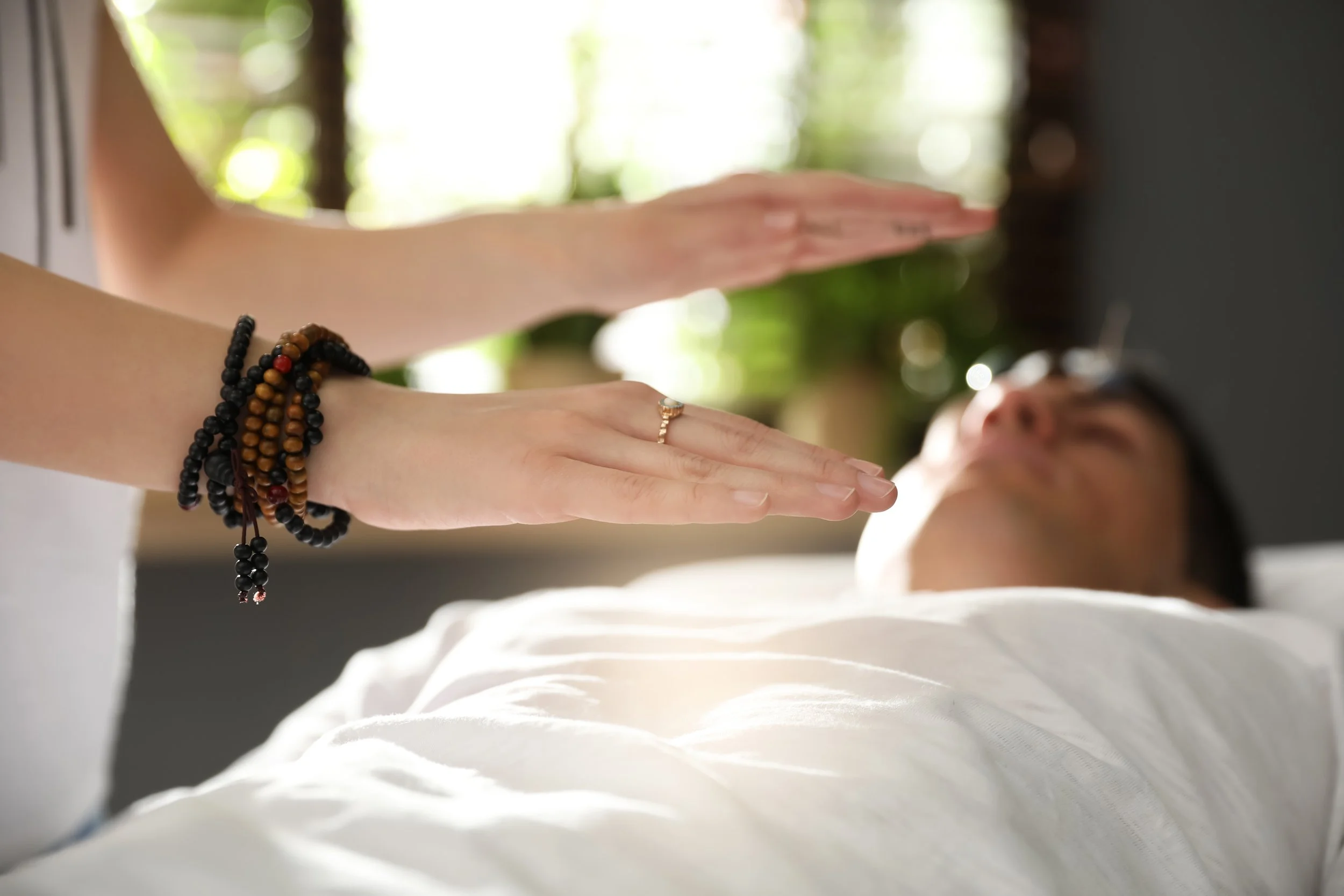 A person lying down on a bed with their eyes closed, receiving a Reiki or energy healing session from a practitioner whose hands are placed above their chest. The practitioner is wearing beaded bracelets and a ring, and the background features a bright, natural setting.