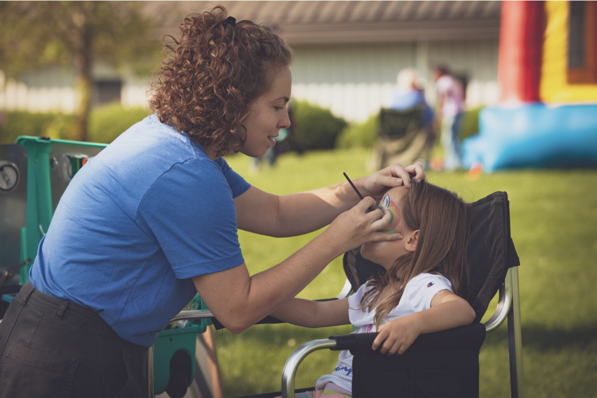 A woman is painting rainbow face paint on a young girl in a wheelchair at an outdoor event.