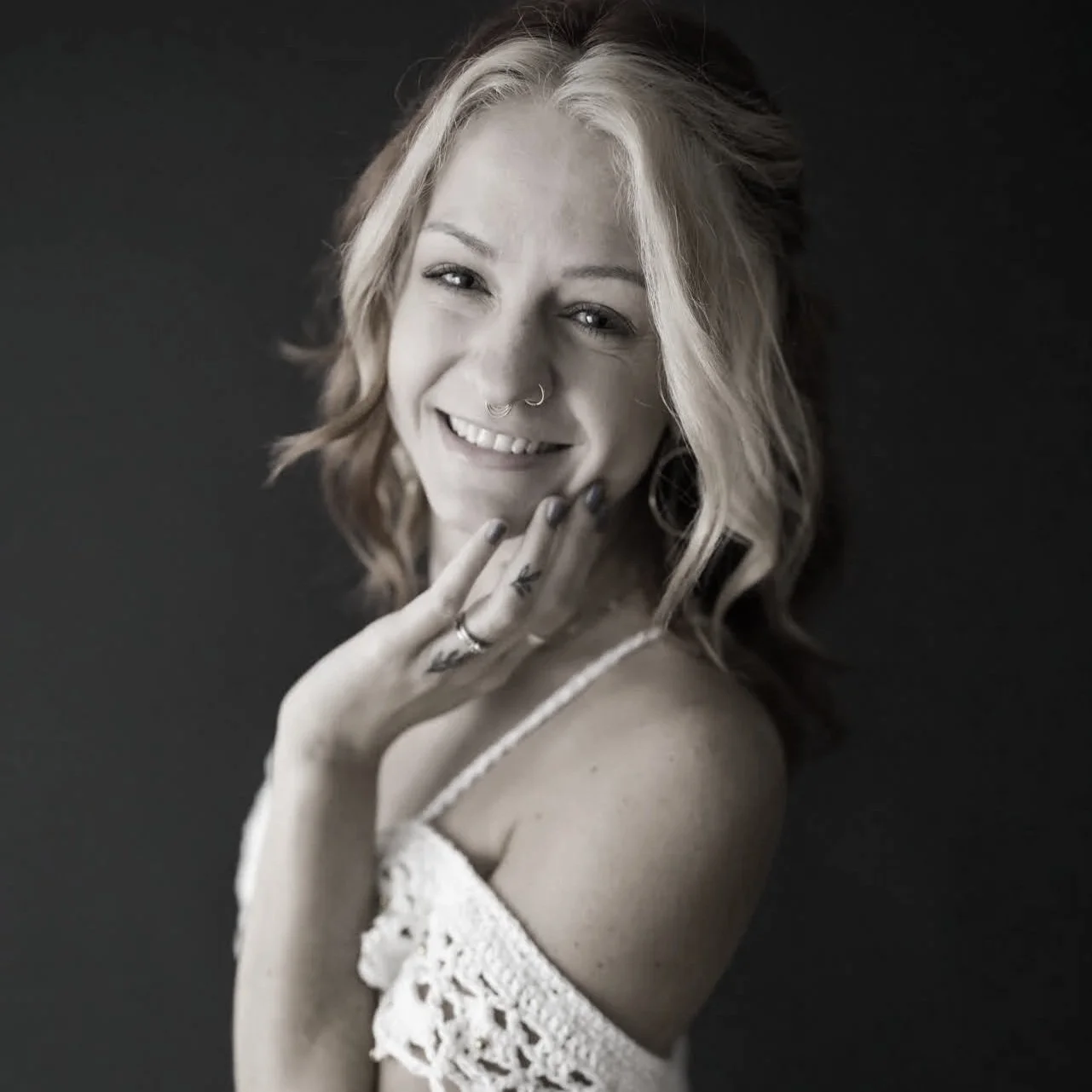 A young woman with wavy hair, smiling and posing in front of a dark background. She has a nose ring, multiple rings on her fingers, and is wearing a lace off-shoulder top.
