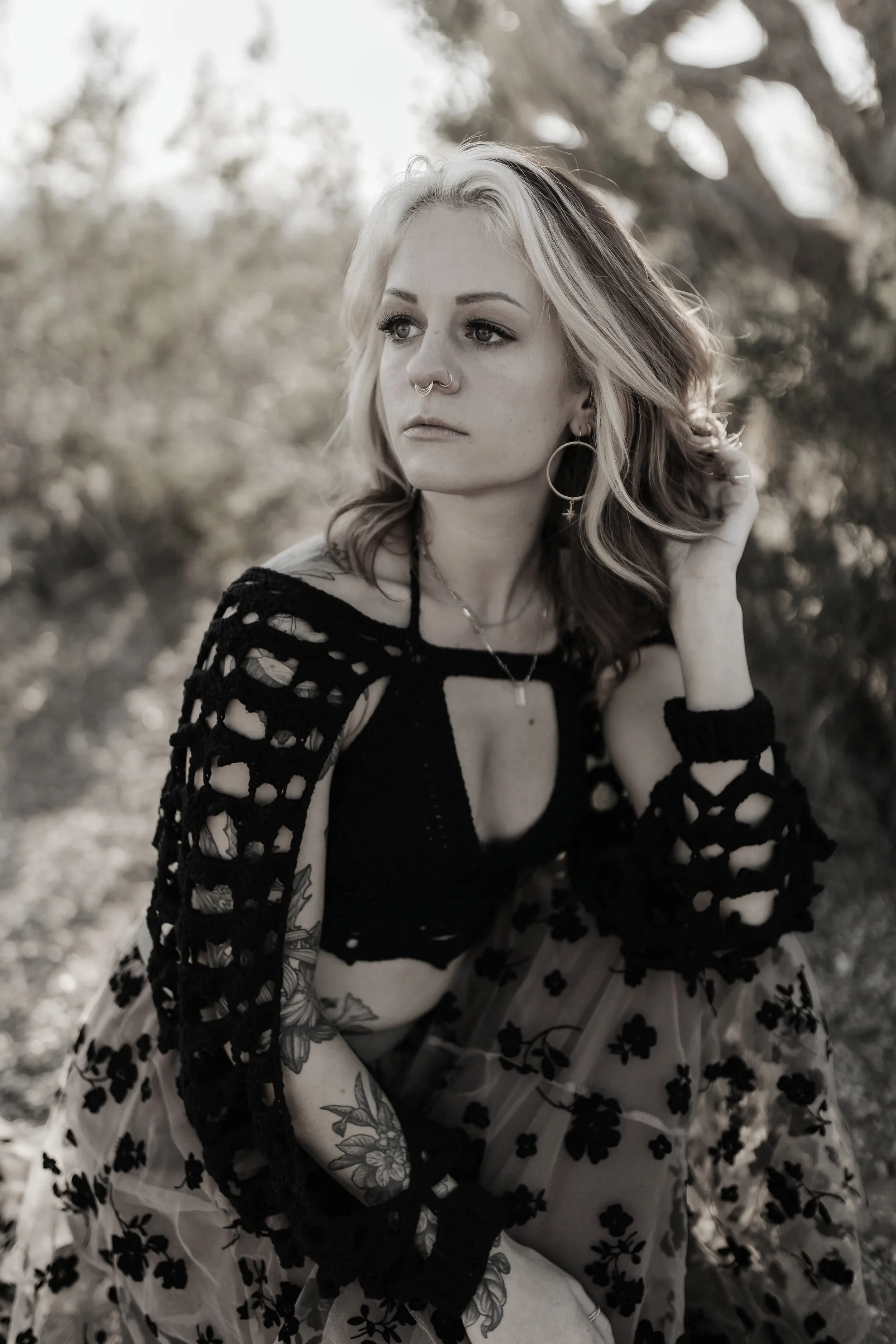 A woman with light hair, tattoos on her arm, wearing a black crochet cover-up over a black top and a floral skirt, sitting outdoors with a thoughtful expression.