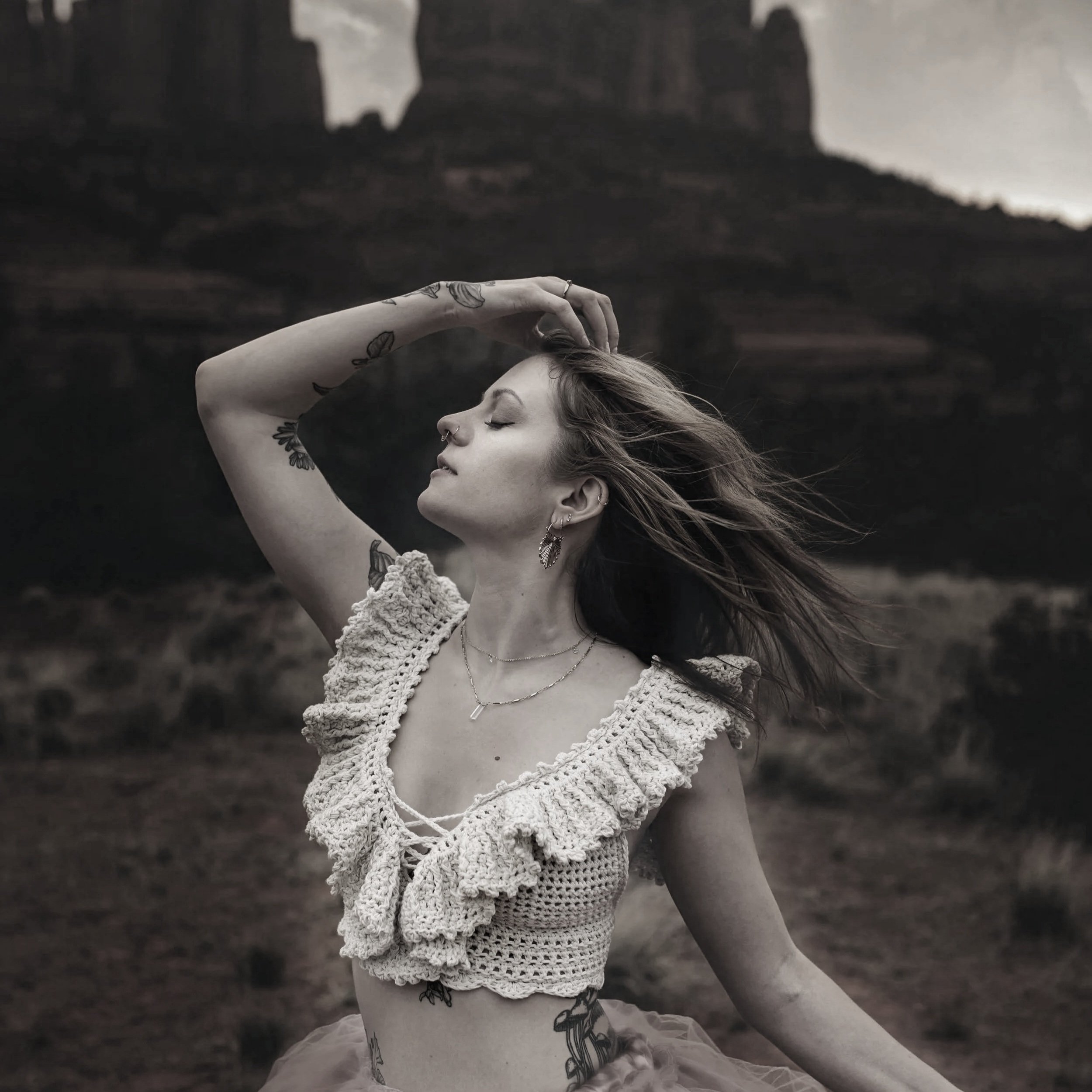 A woman with tattoos, earrings, and piercings, in a crocheted top, stands outdoors with her eyes closed, wind blowing her hair, holding her head with one hand, in a blurred natural landscape background.
