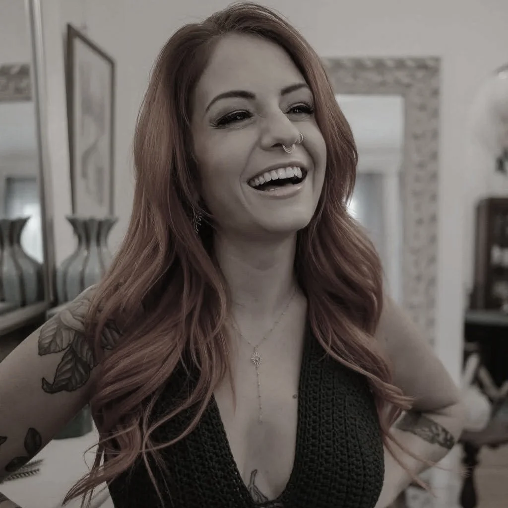 A woman with long wavy hair, partially dyed pink, is smiling in a black sleeveless top, wearing a necklace, earrings, and a septum piercing, in a home setting with vases and picture frames in the background.