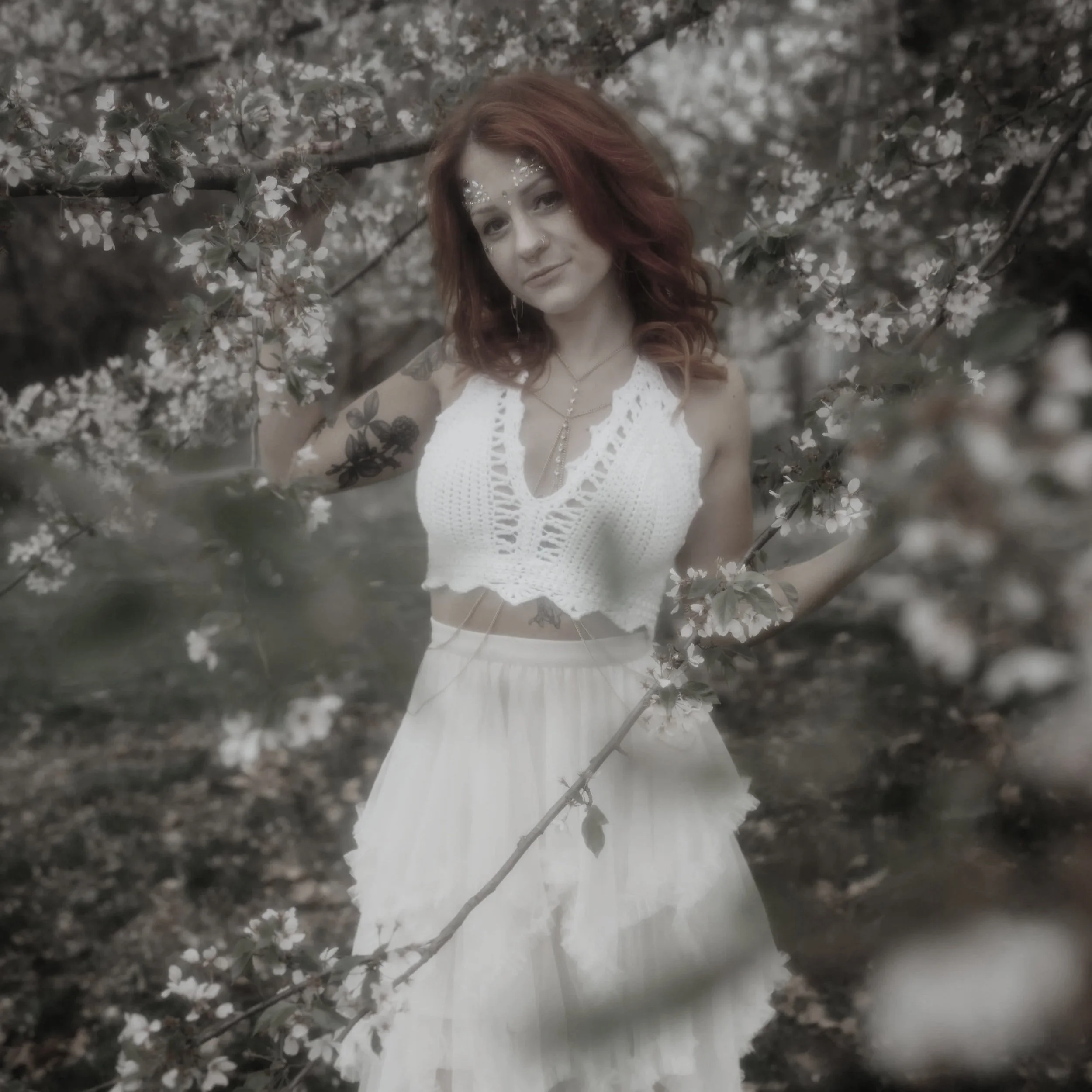 A young woman with red hair, tattoos, and face decorations is standing among blooming branches, wearing a white crochet top and a flowing skirt.