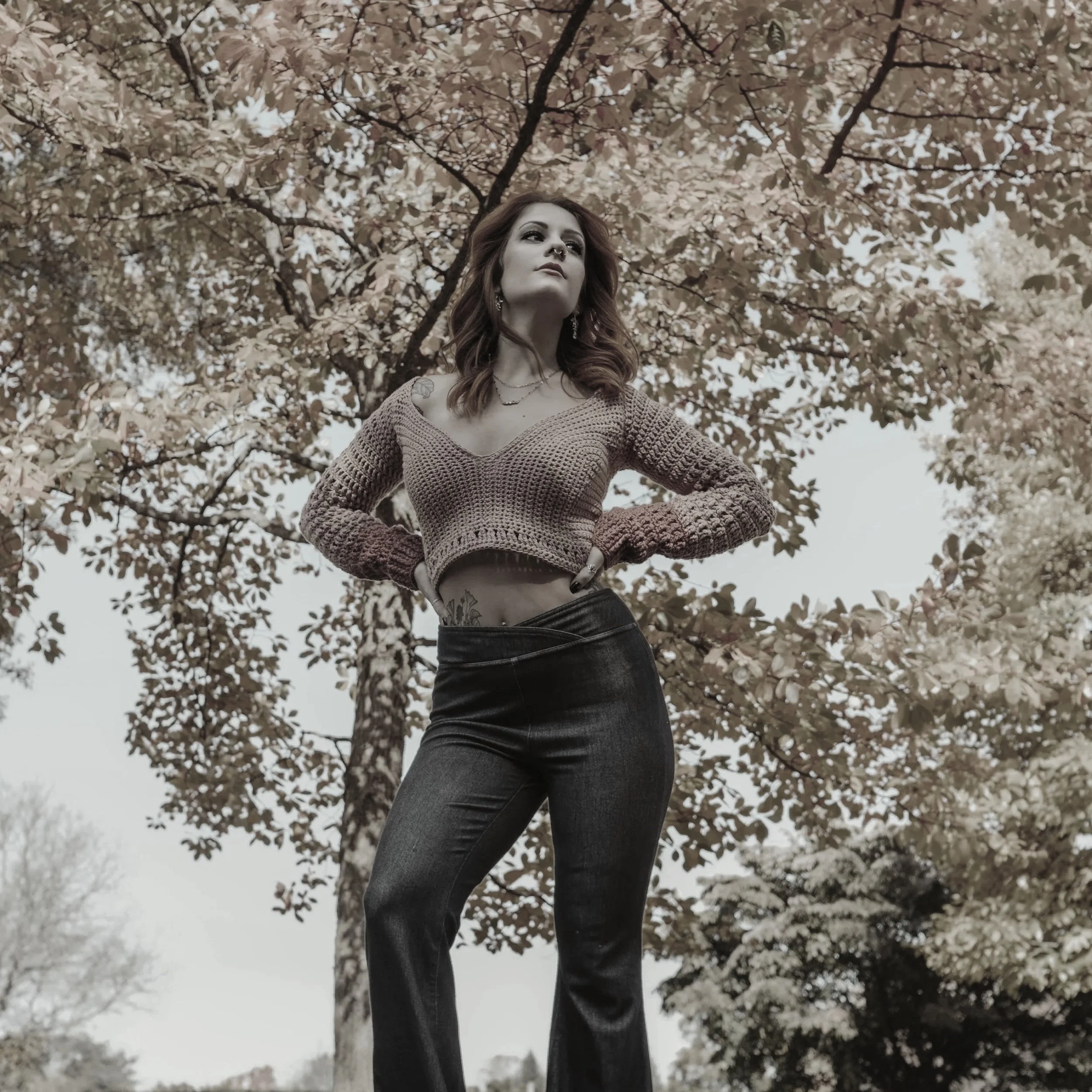 A woman standing outdoors beneath a tree with pink leaves, wearing a crochet crop top and black jeans, with her hands on her hips.