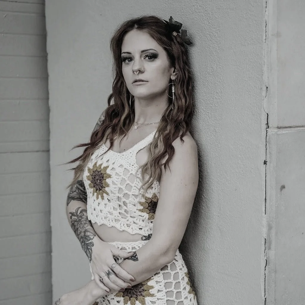 A young woman with long wavy hair, tattoos, and jewelry, standing against a textured wall, wearing a crochet top and skirt with floral patterns and looking directly at the camera.