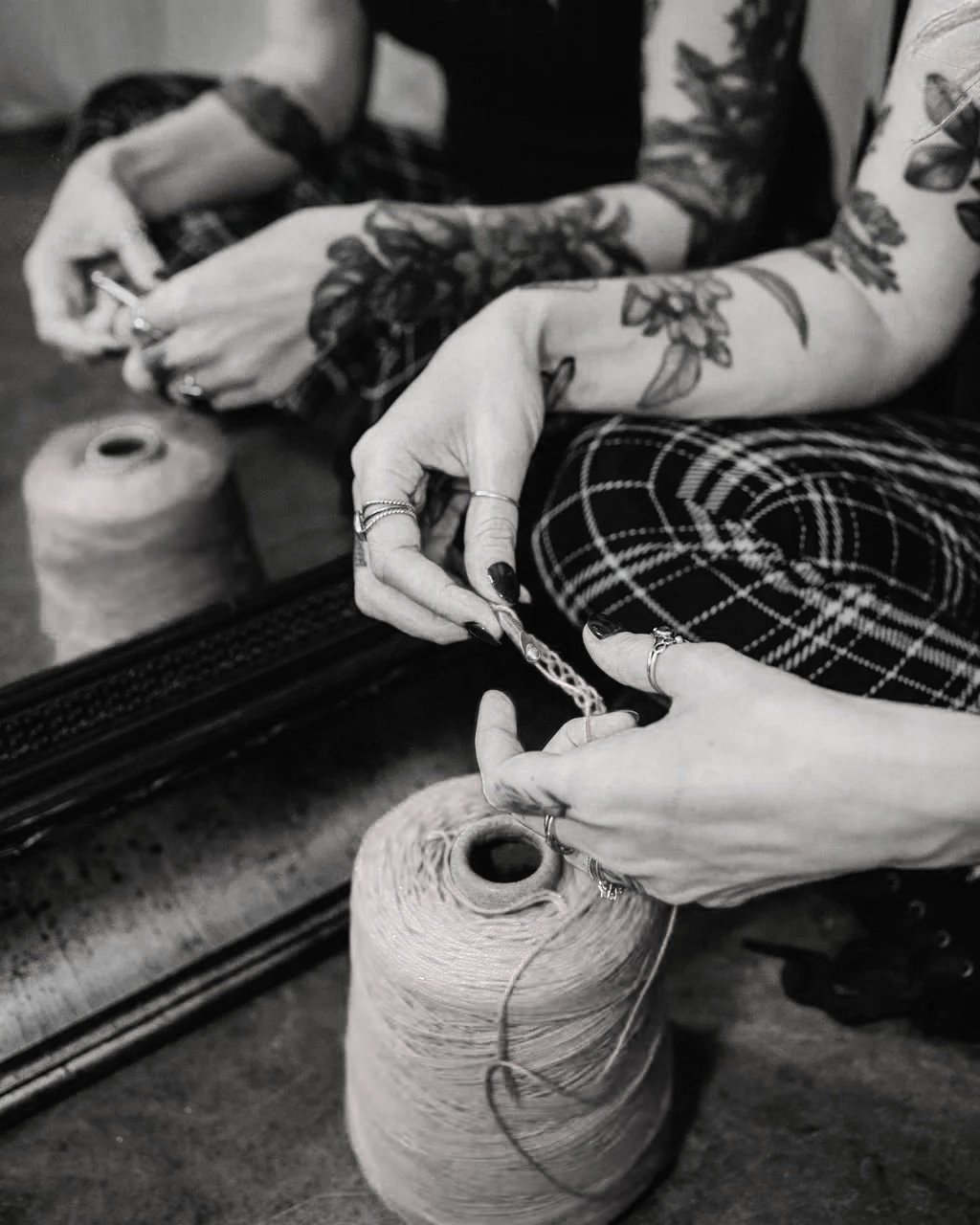 Close-up of tattooed hands working with thread and needle, sitting beside large spool of thread.